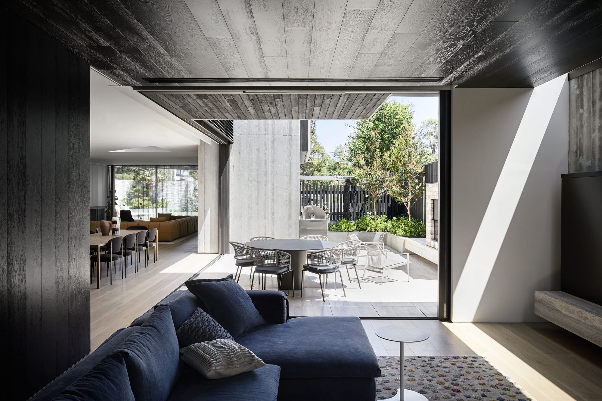 The Split Home by Seidler Group. Copyright of Seidler Group. Living room opening onto external courtyard with outdoor dining setting. Navy couch inside and black timber walls and ceilings.