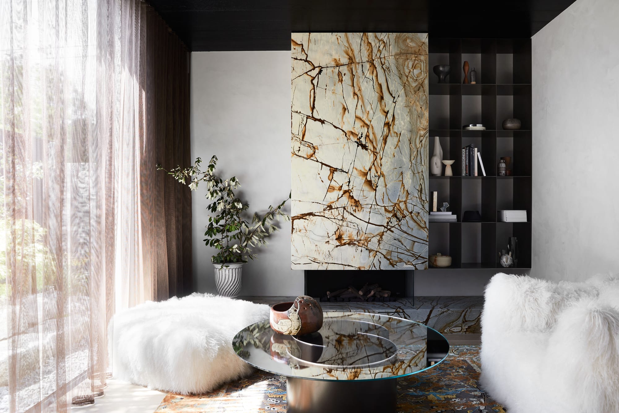 The Split Home by Seidler Group. Copyright of Seidler Group. Small sitting room with marble fireplace. Fluffy, white stools arranged around mirrored circle coffee table. 