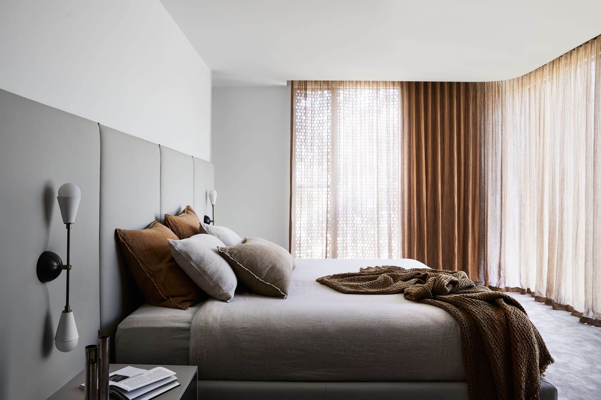 The Split Home by Seidler Group. Copyright of Seidler Group. Residential bedroom with floor to ceiling windows covered with orange sheer curtains. Grey upholstered bedhead runs length of wall behind bed with grey linen. 
