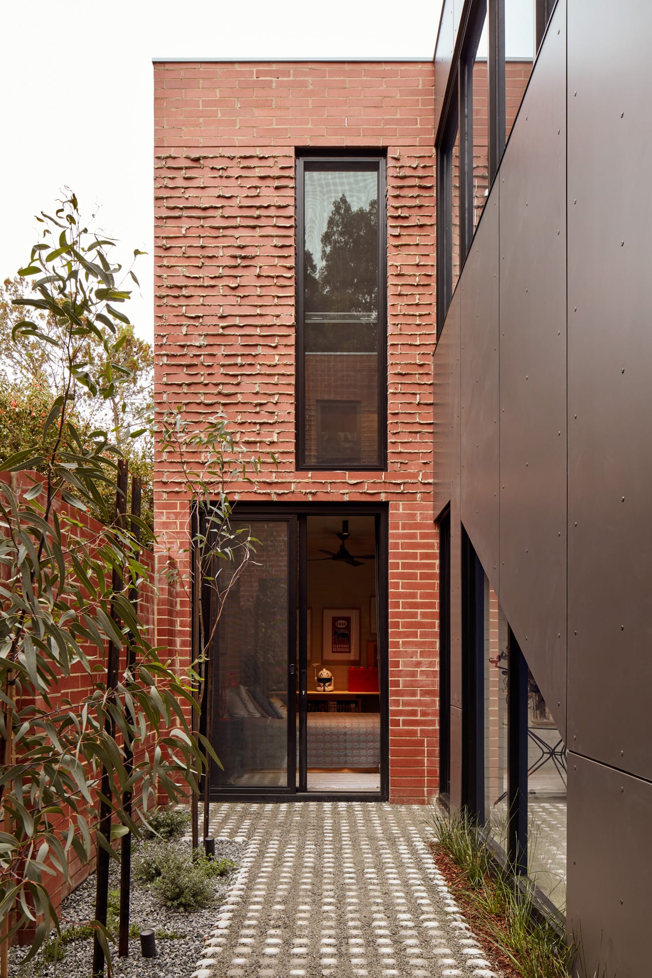 Brick House by Studio Roam. Photography by Jack Lovel. Double storey brick facade with block windows and door. Black metal wall to right. 