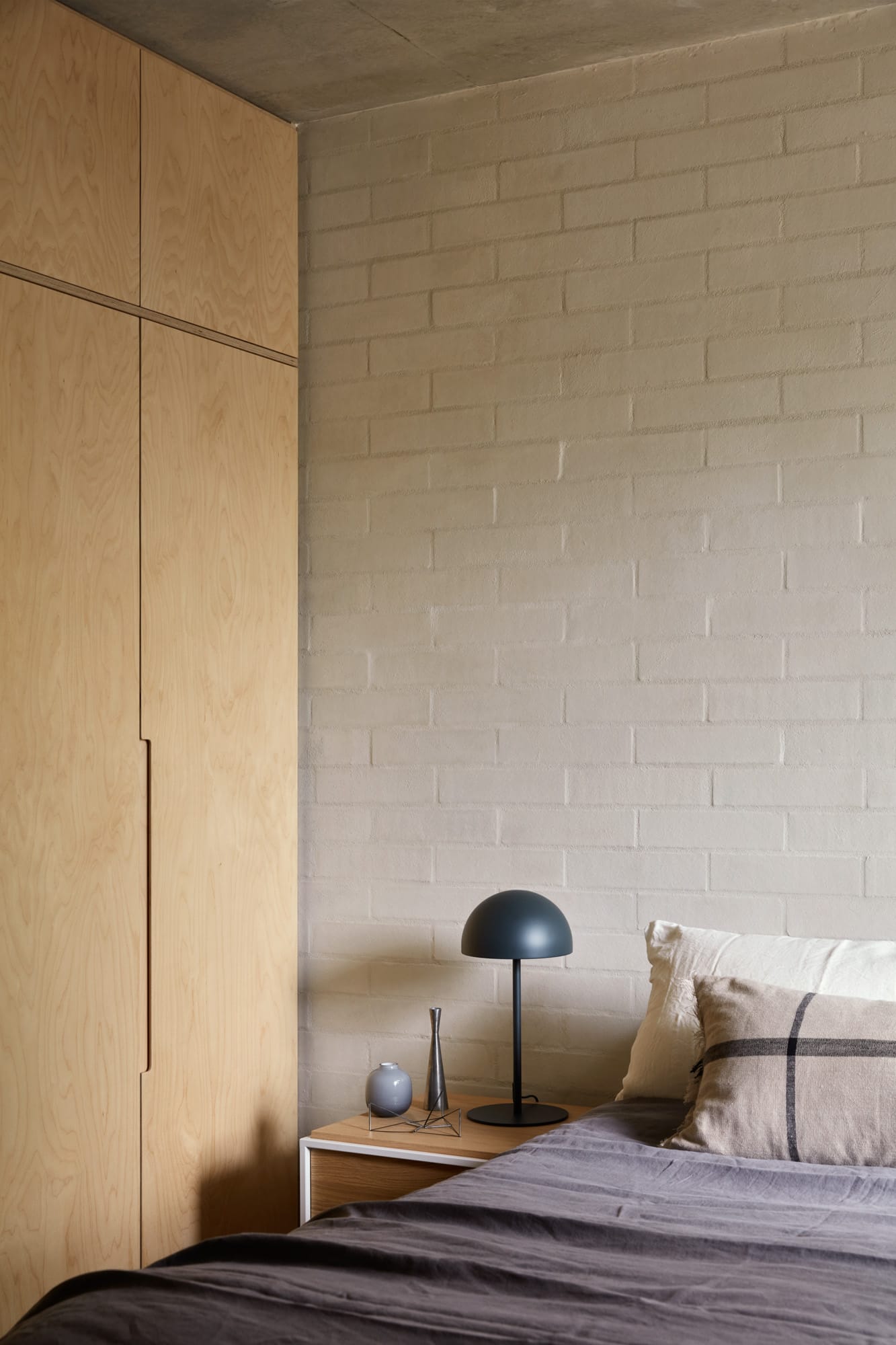  Brick House by Studio Roam. Photography by Jack Lovel. Bedroom with plywood wardrobes and white brick walls. Grey linen on bed. Timber bedside table with black lamp. 