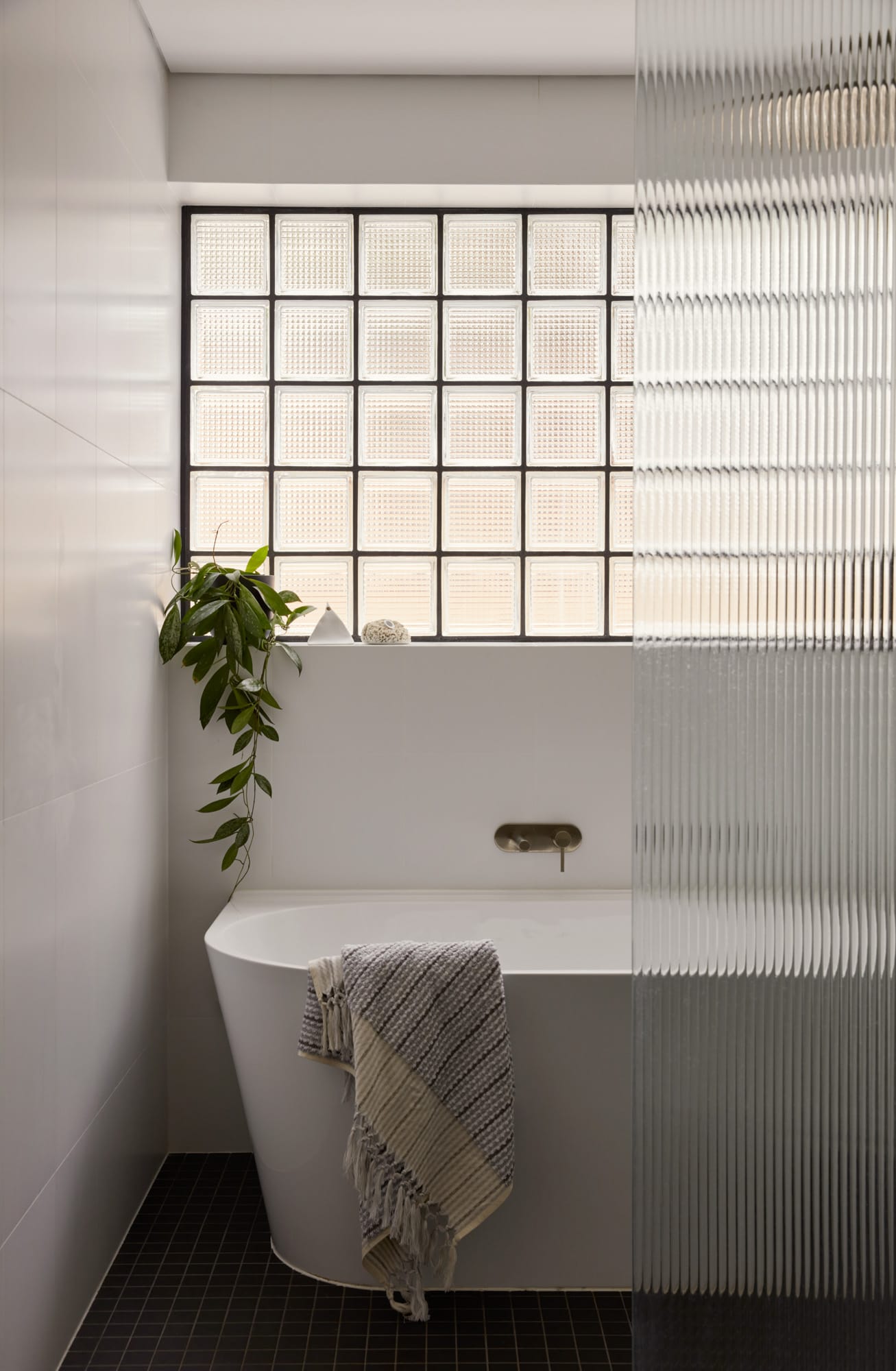 Brick House by Studio Roam. Photography by Jack Lovel. Bathroom with freestanding white tub. Fluted glass shower screen. Glass block window. Black floor tiles. 