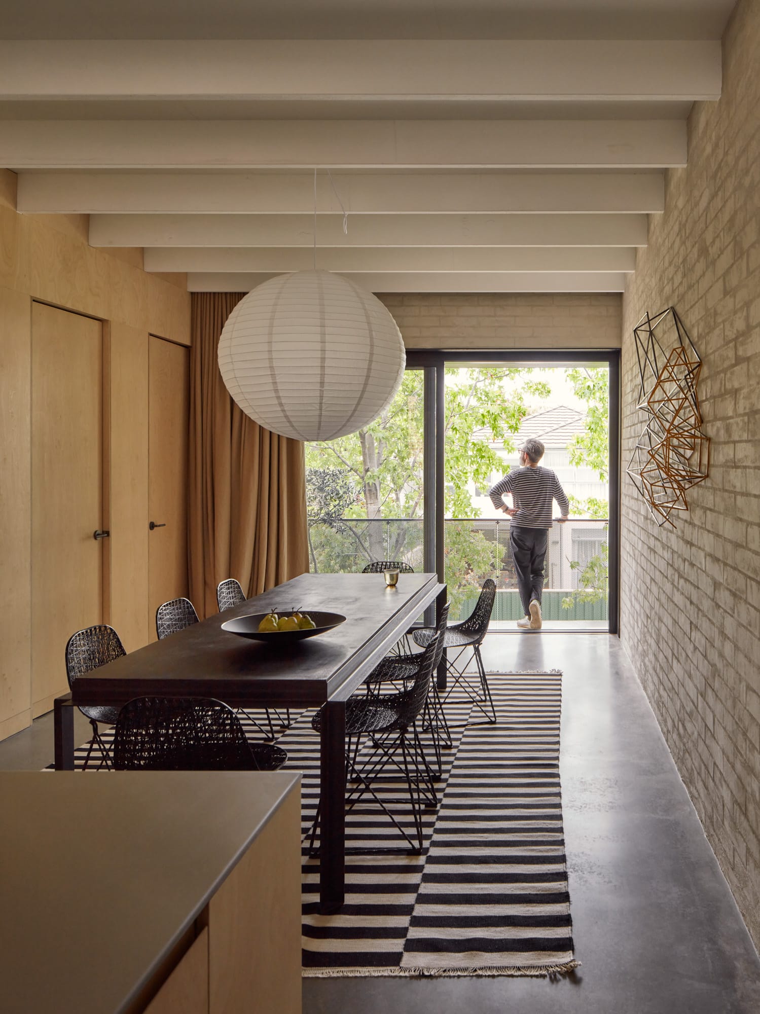 Brick House by Studio Roam. Photography by Jack Lovel. Large black dining table with black dining chairs. Polished concrete floors. Brick wall to right, plywood wall to left. Dining space extending out to balcony overlooking street. 