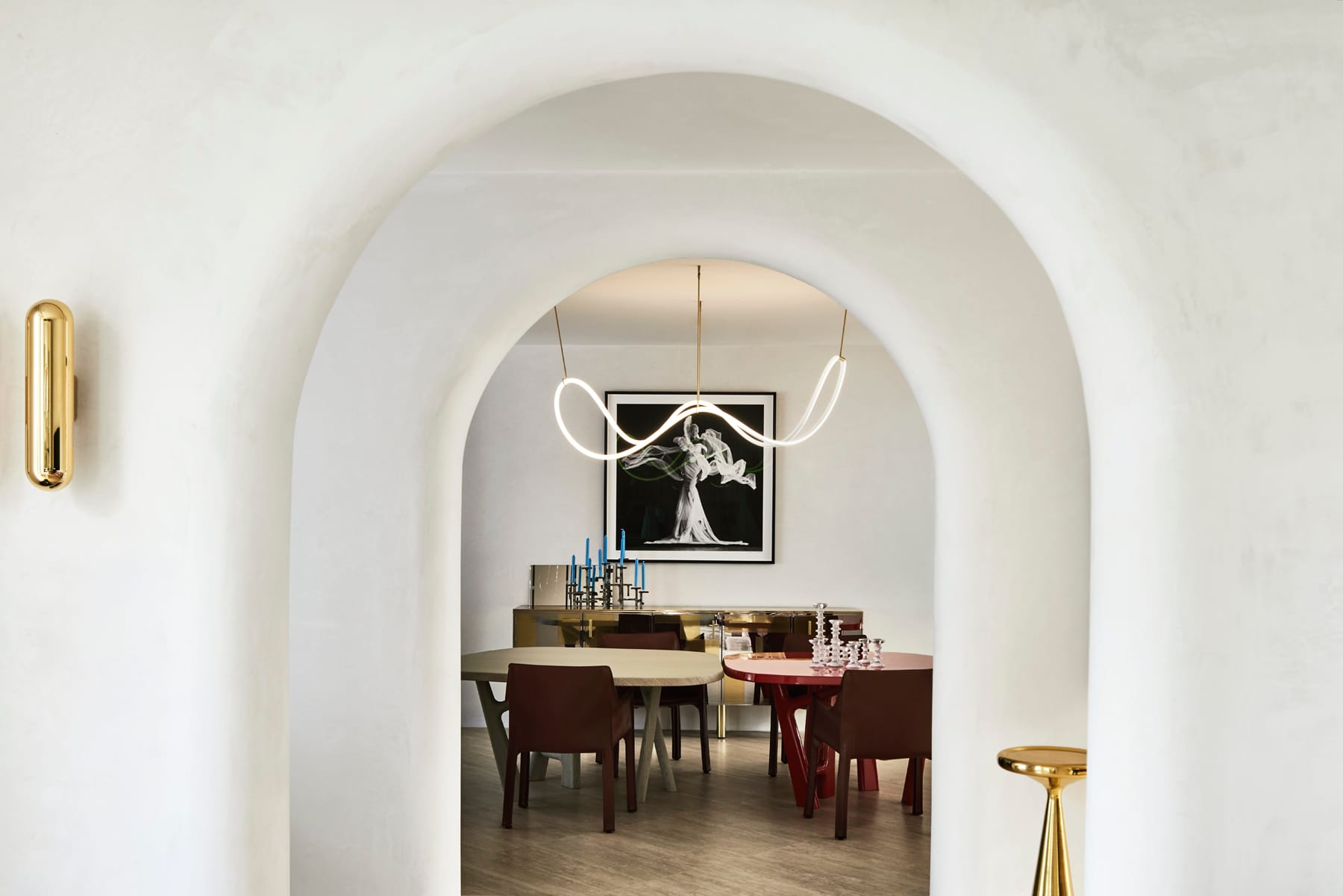 Toorak House 2 by K.P.D.O. Photography by Sharyn Cairns. Multiple archways framing dining room. Red and white table with red dining chairs. Abstract pendant lighting. Gold furniture scattered throughout. 