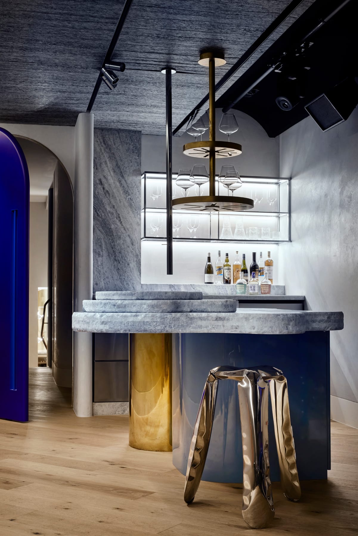 Toorak House 2 by K.P.D.O. Photography by Sharyn Cairns. Navy blue bar with layered stone benchtop. Bronze leg detailing and gold abstract bar stool. Electric blue door to left. 
