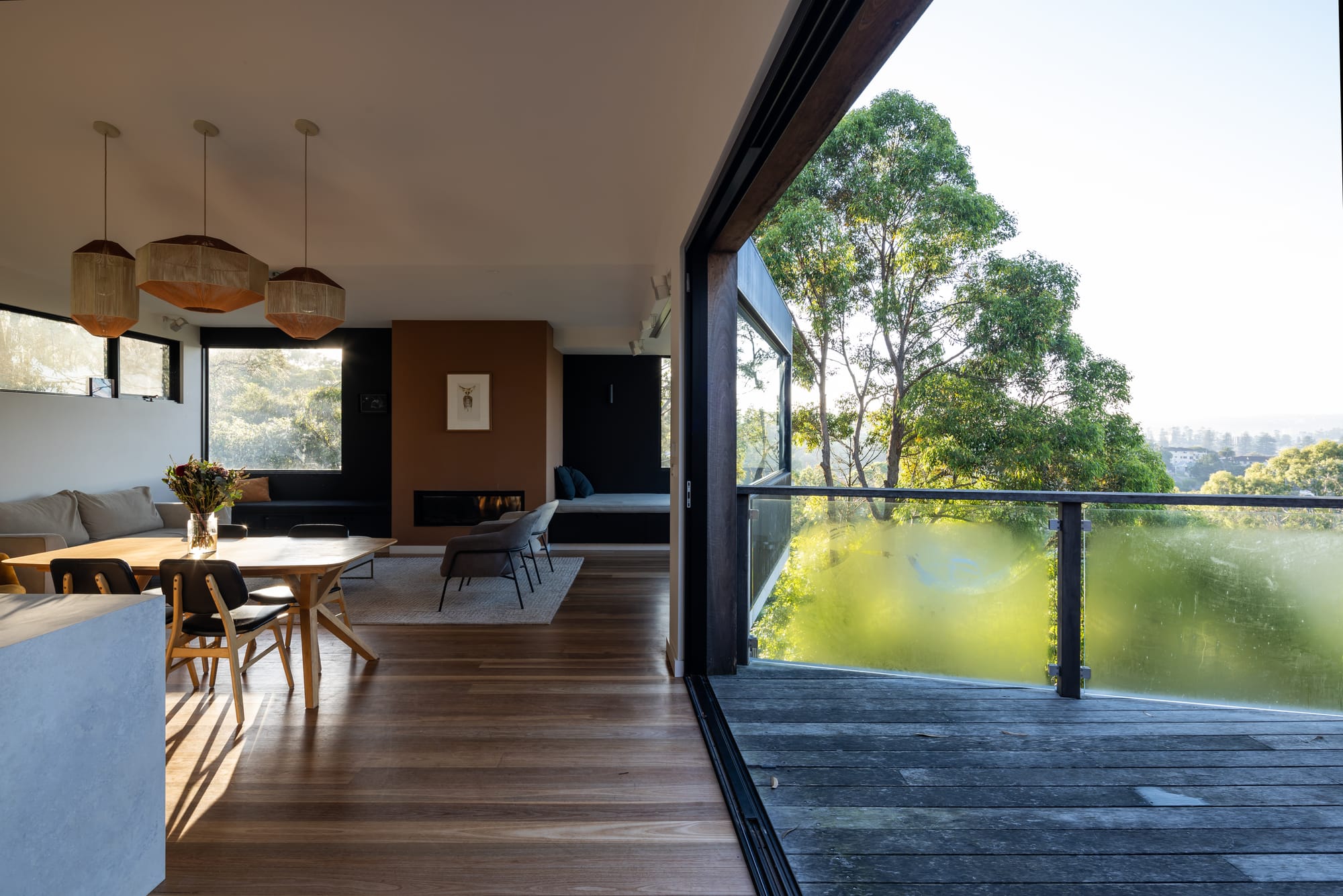 Tree House by North By North. Photography by Simon Whitbread. Open plan kitchen, living dining space extending onto timber deck. Glass fencing on deck overlooking green views. 