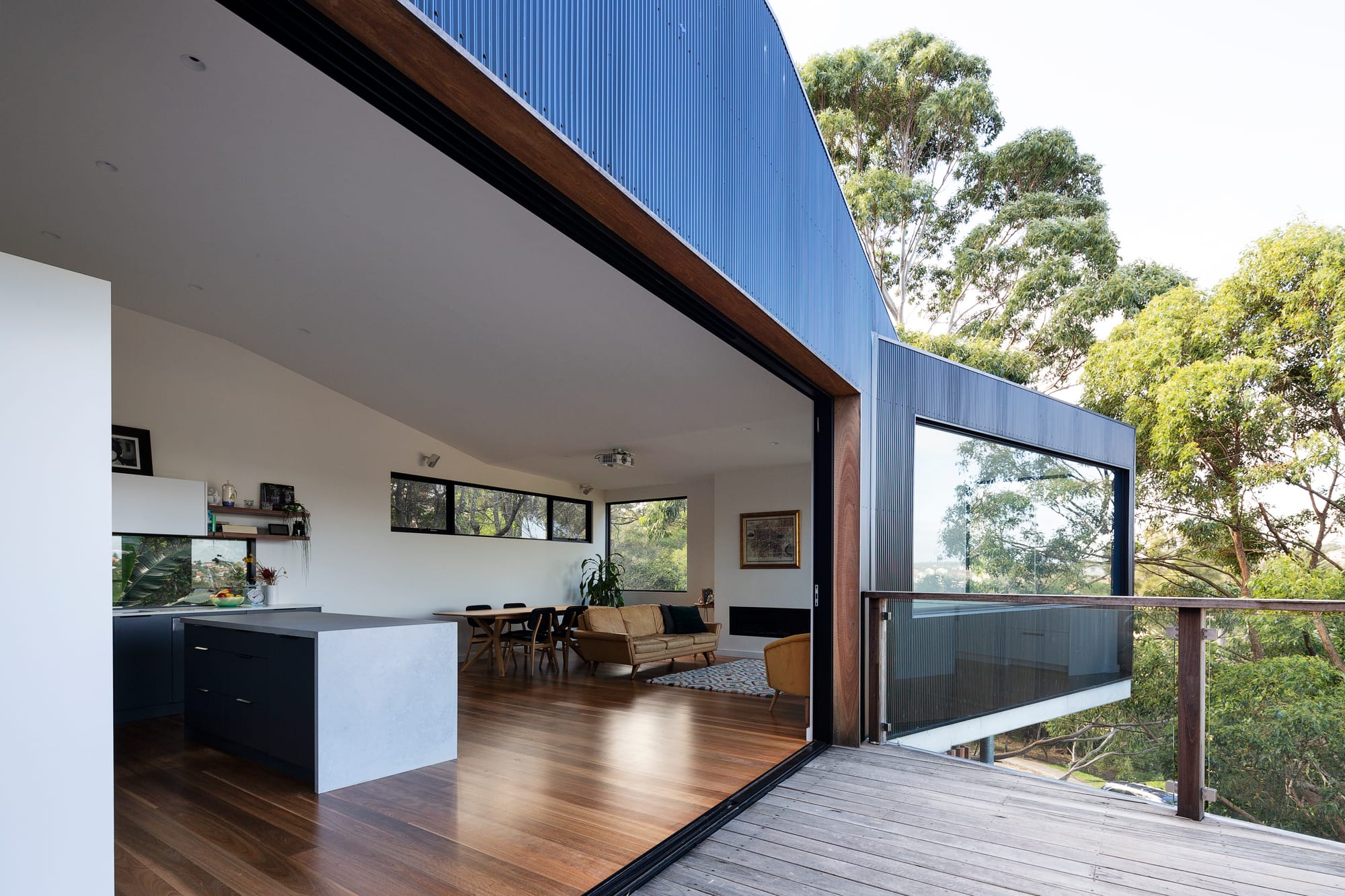 Tree House by North By North. Photography by Simon Whitbread. Open plan living space extending onto timber deck. Views of trees. Black kitchen cabinetry.