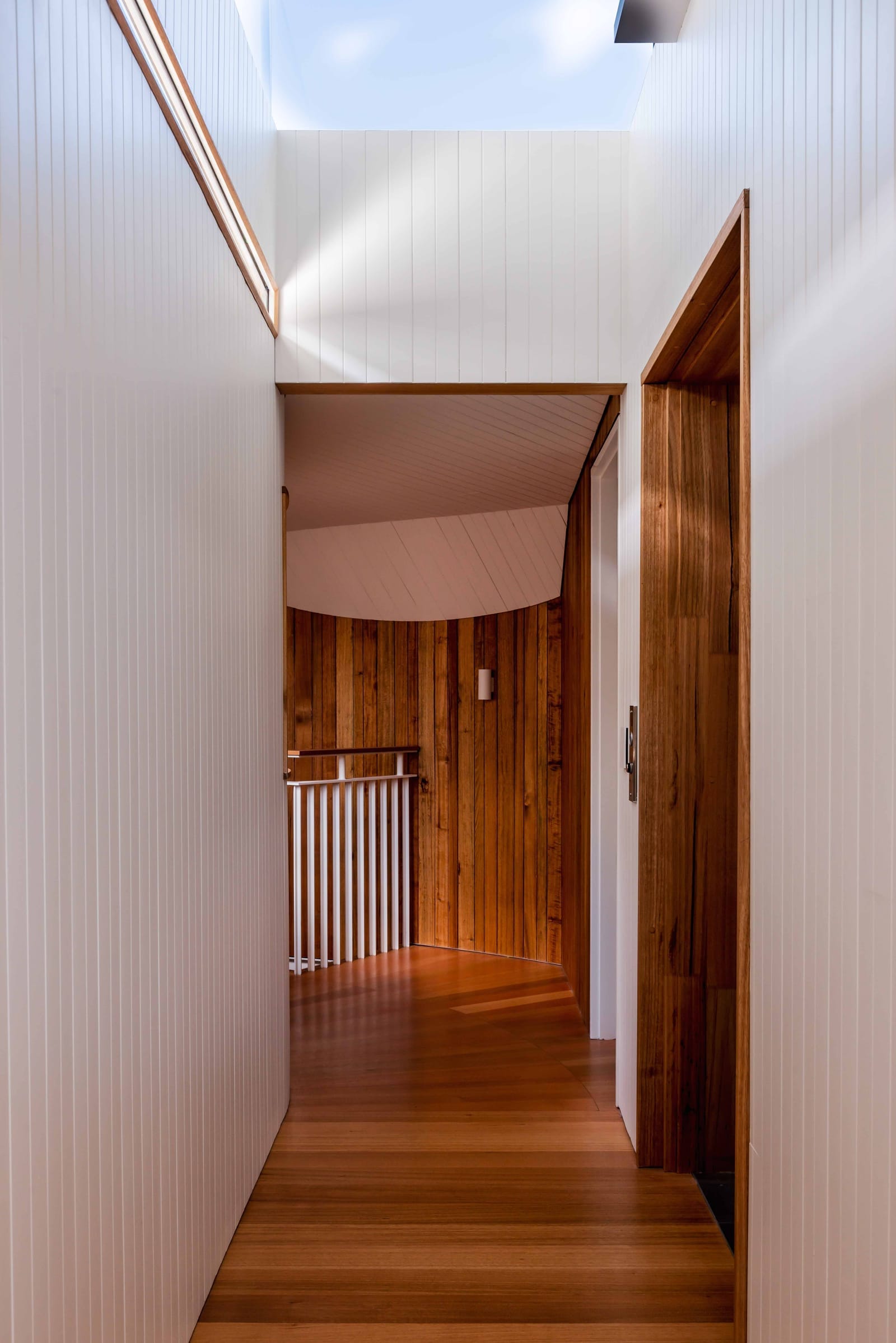 Wattle Bird House by Flett Architecture. Photography by Matt Sansom. Hallway with polished timber floors and white timber clad walls. White metal staircase balustrade at end of hallway. 