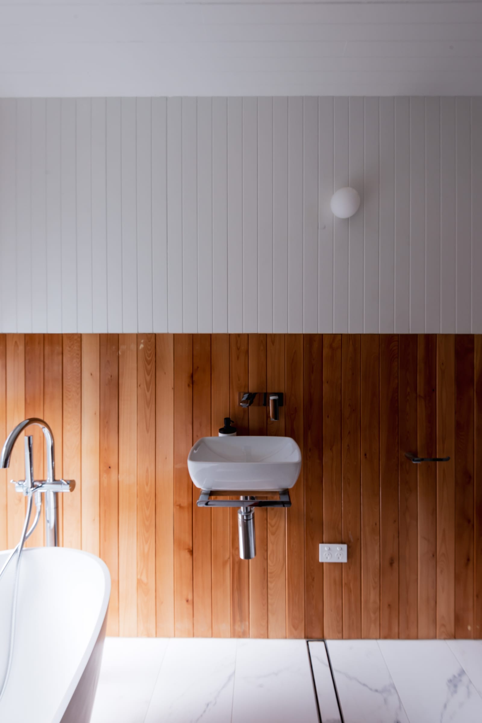 Wattle Bird House by Flett Architecture. Photography by Matt Sansom. White wall-mounted sink on timber paneled bathroom wall. Marble tiled floor. White freestanding bath to left.  