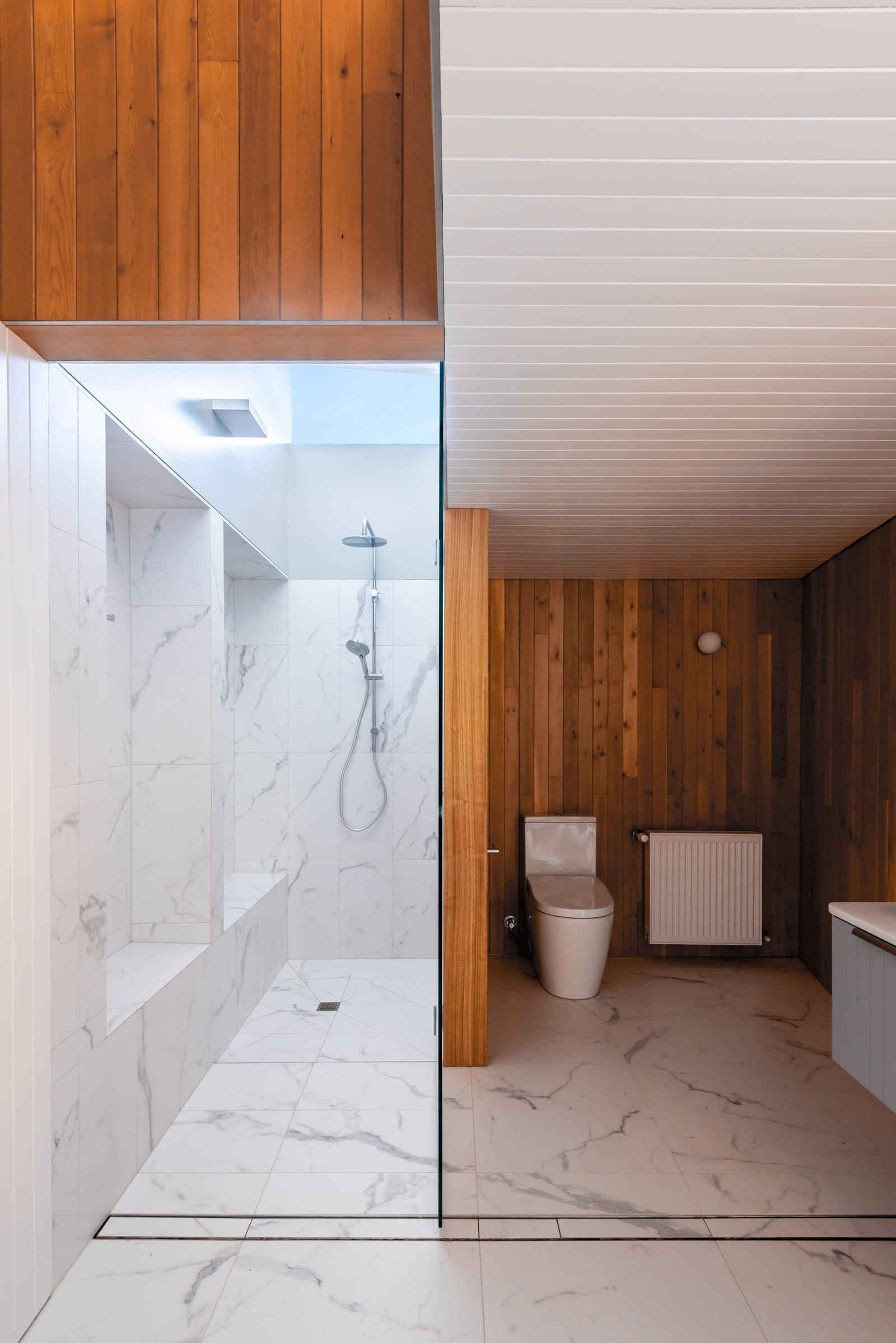 Wattle Bird House by Flett Architecture. Photography by Matt Sansom. Bathroom with marble floors and walls. Timber paneled walls around toilet and on ceiling. Ceiling light in shower. 