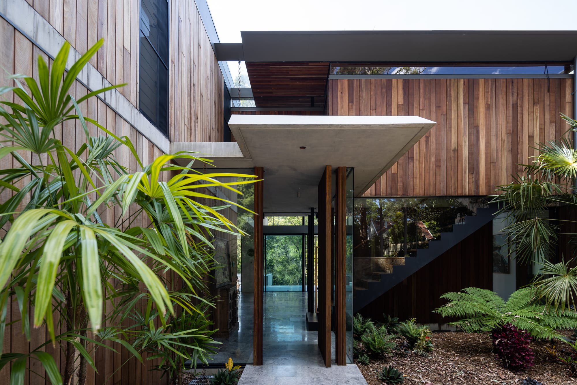 Mortlock Timbers Series: Fox Valley House. Photography by Simon Whitbread. Entrance to double storey residential home. Timber clad exterior walls and enclosed glass entryway. Garden with native plants. 