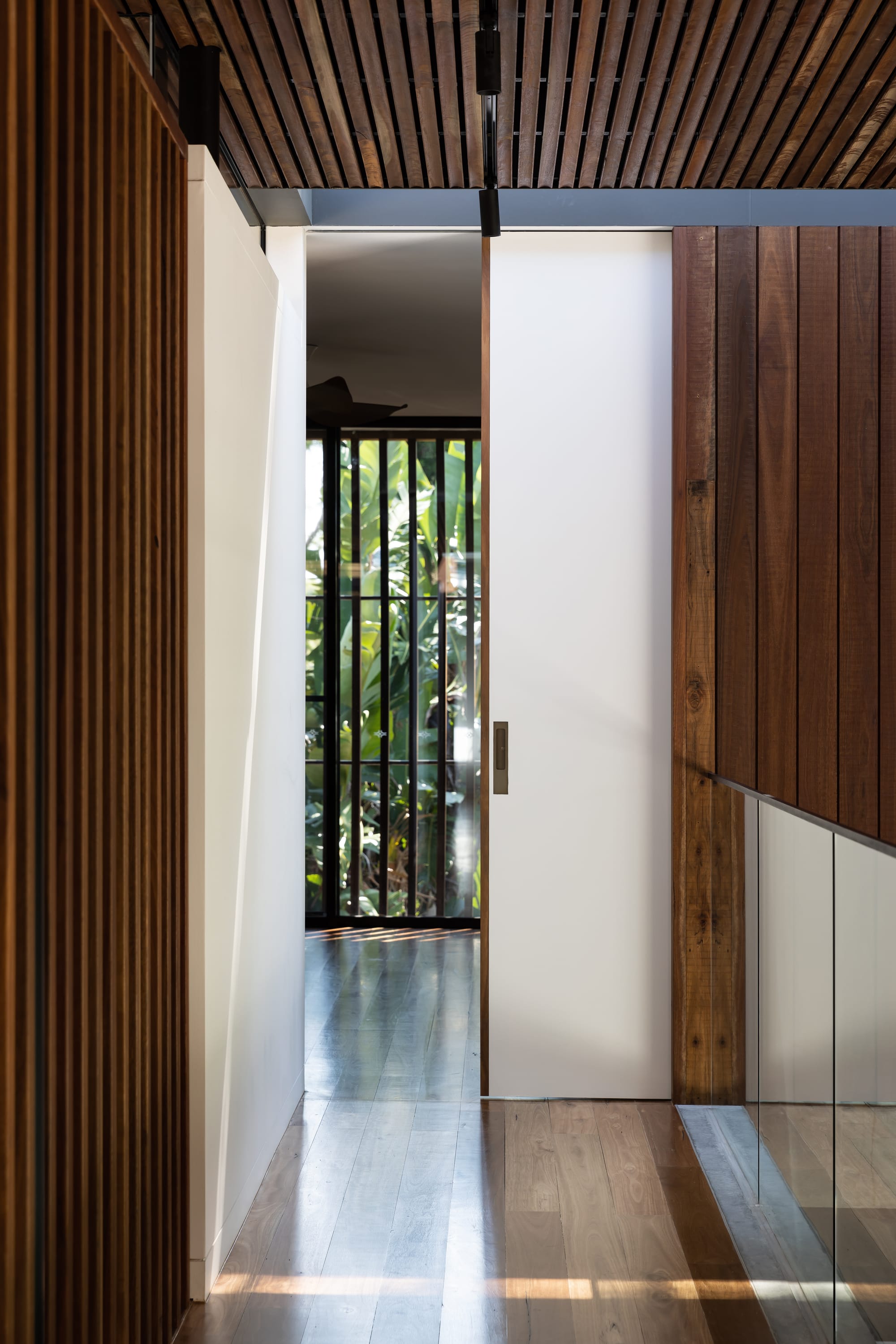 Mortlock Timbers Series: Fox Valley House. Photography by Simon Whitbread. Hallway with timber flooring, walls and ceiling. White door opening into room with garden views. Glass balcony fence.
