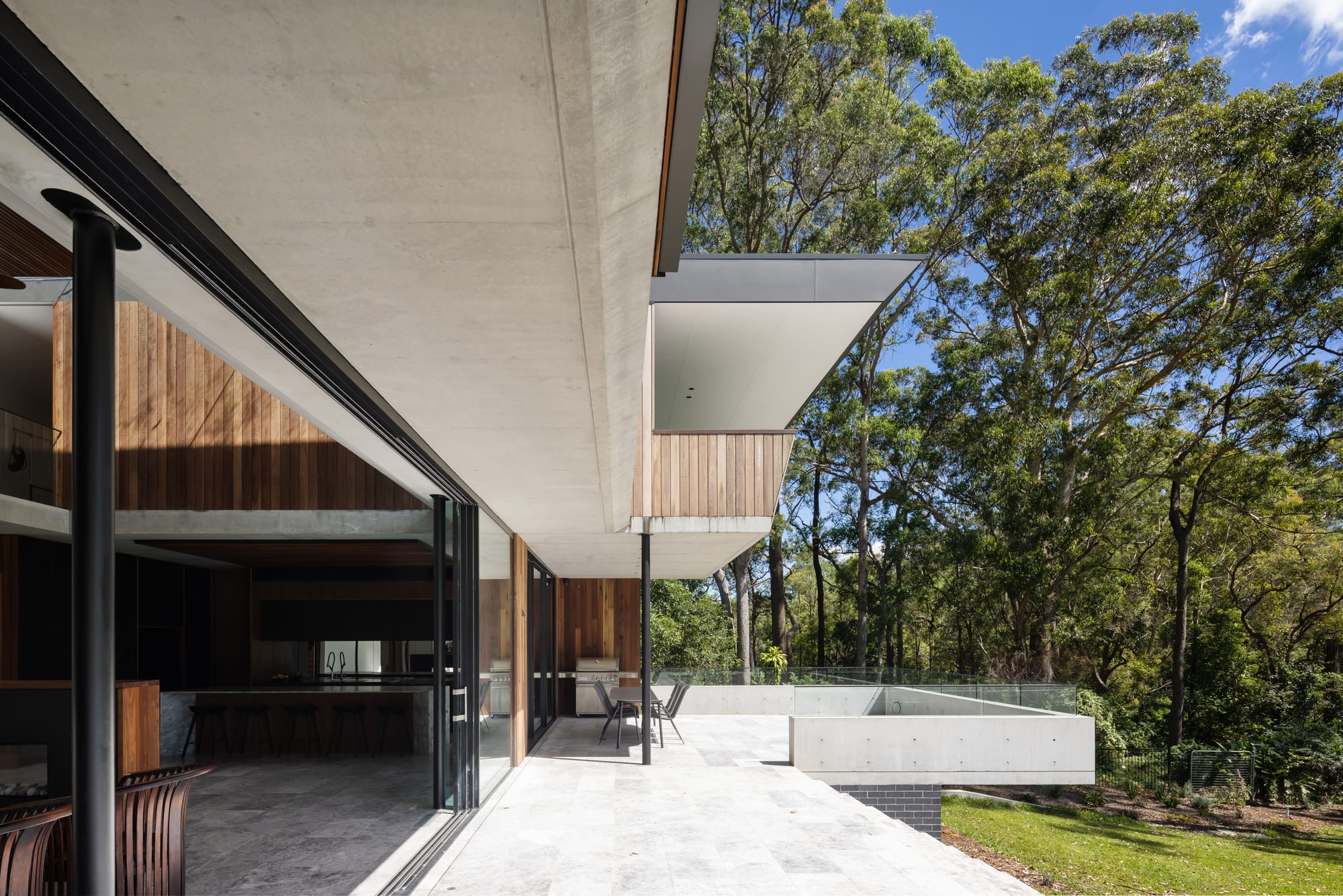 Mortlock Timbers Series: Fox Valley House. Photography by Simon Whitbread. Undercover patio running length of house with verandah outcropping. Concrete floor and ceiling. Grassy hill to right, interior living space to left. 