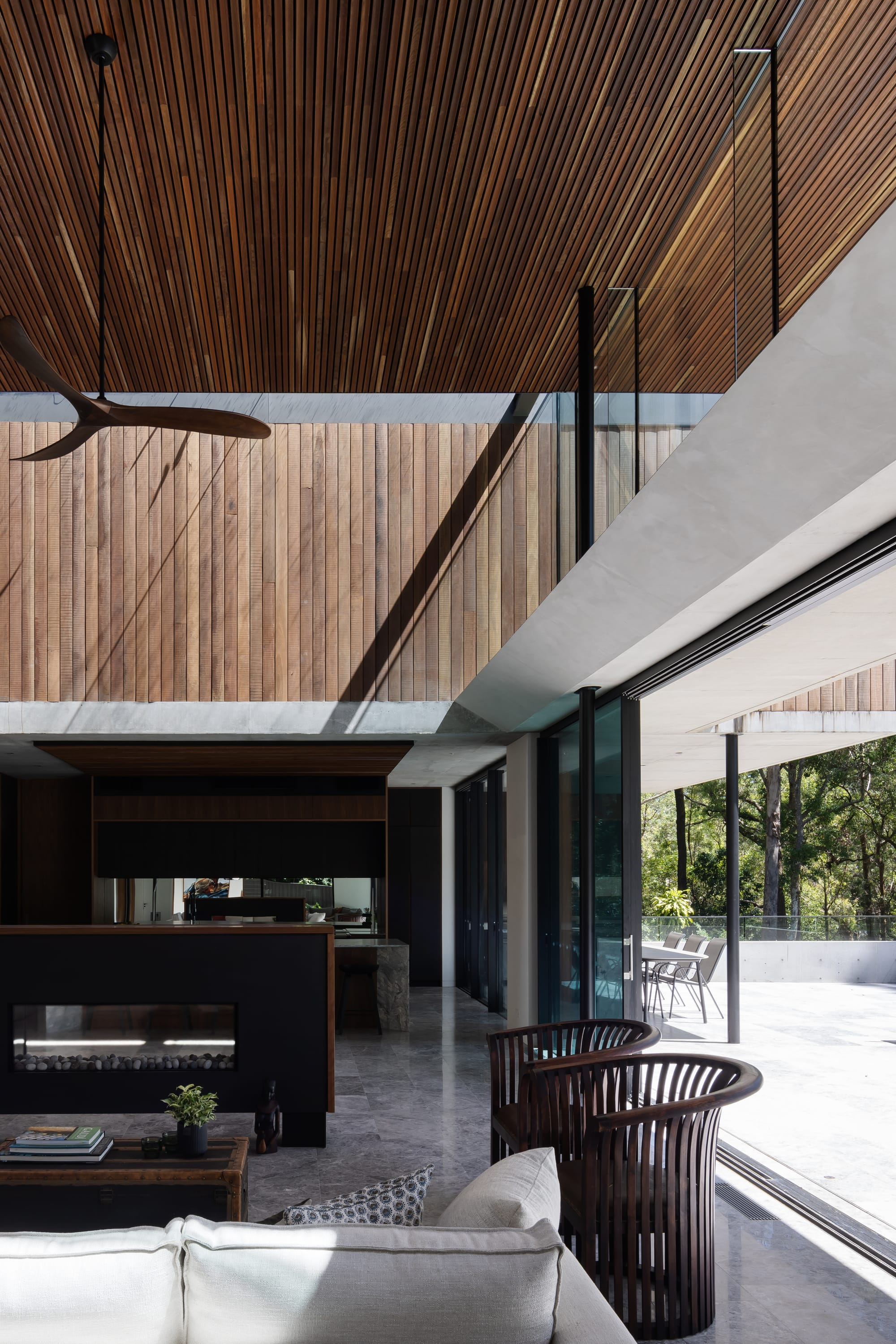 Mortlock Timbers Series: Fox Valley House. Photography by Simon Whitbread. Double storey interior living space with polished stone floors and timber clad walls and ceiling on second floor. Timber armchairs and white lounge in foreground.