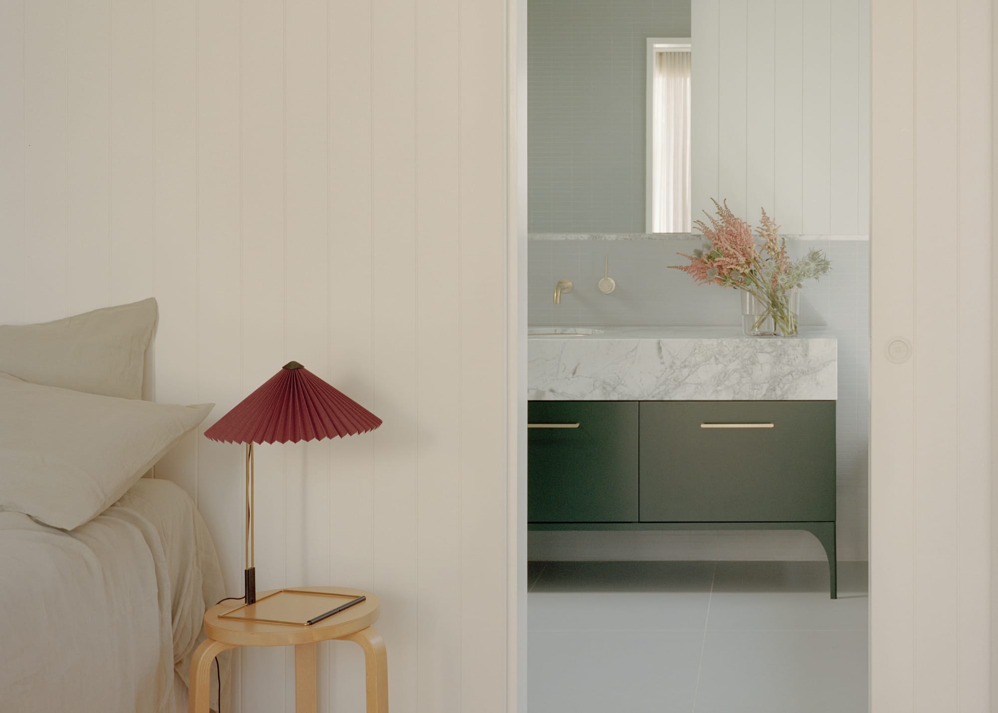 Gable Park by Weaver+Co Architects. Photography by Tasha Tylee. White timber clad walls in bedroom. Bedside table with red lamp. Ensuite with dark green and marble counter. 