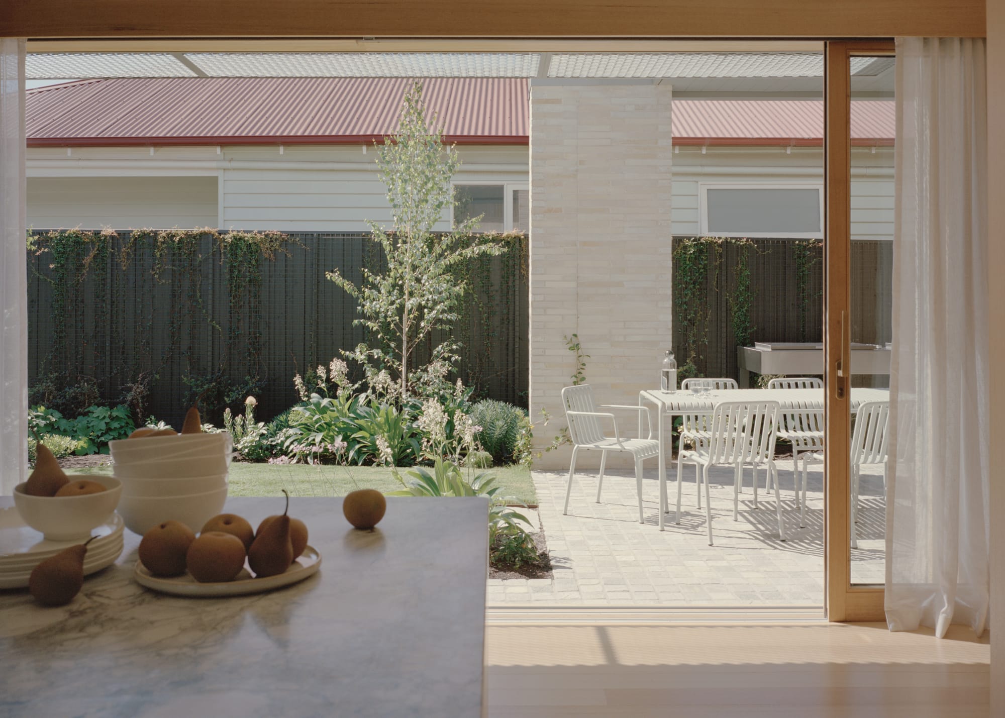 Gable Park by Weaver+Co Architects. Photography by Tasha Tylee. View to patio and courtyard through sliding timber framed doors in kitchen. White outdoor dining setting and green grass.