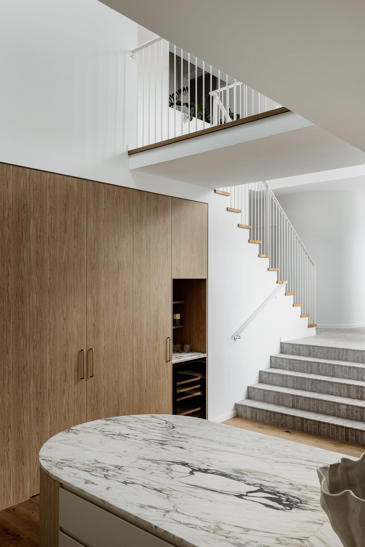 Hazlewood House by Favell Architects. Photography by Andy Macpherson. Kitchen with timber floors and timber cabinetry. White marble countertop. Grey tiled steps leading away from kitchen, white and timber staircase to left of image. 