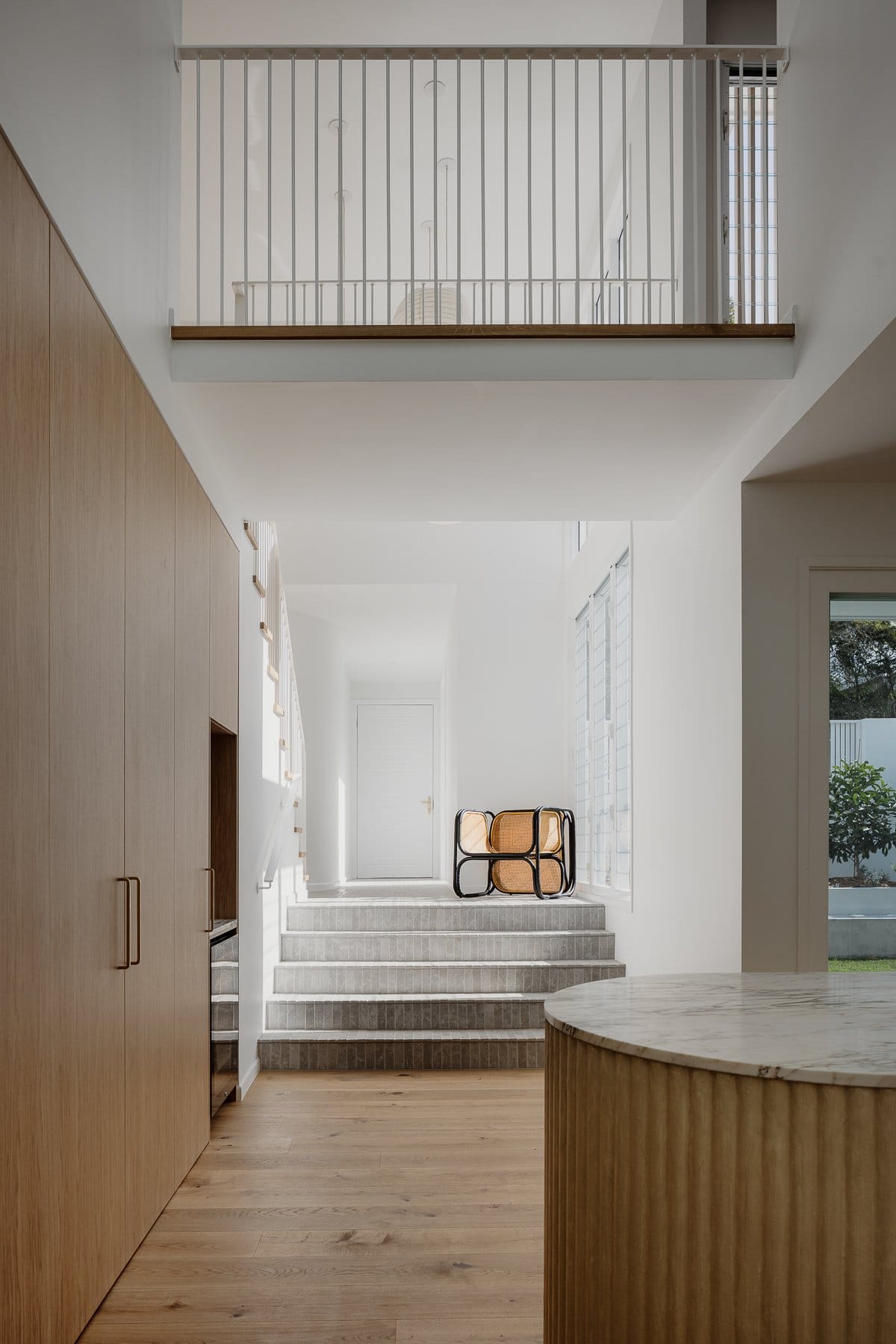 Hazlewood House by Favell Architects. Photography by Andy Macpherson. Tiled steps leading to white hallway and away from kitchen. Timber floors and cabinetry in kitchen in forefront. Black and wicker chair in centre of image. 