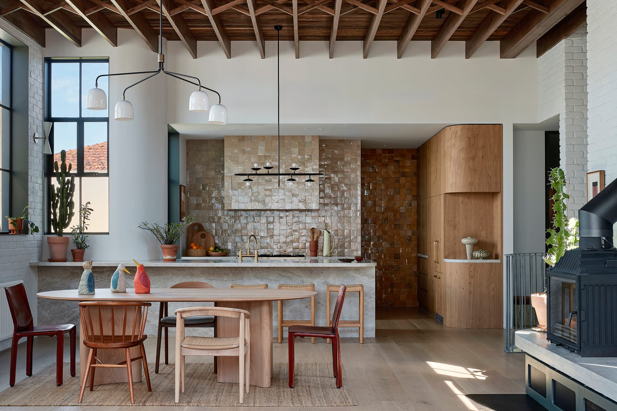 Fabrica by mcmahon and nerlich. Photography by Shannon McGrath. Open plan dining and kitchen with exposed ceiling beams and white plaster walls. Timber dining table with eclectic chairs. Beige tiles on wall in kitchen.