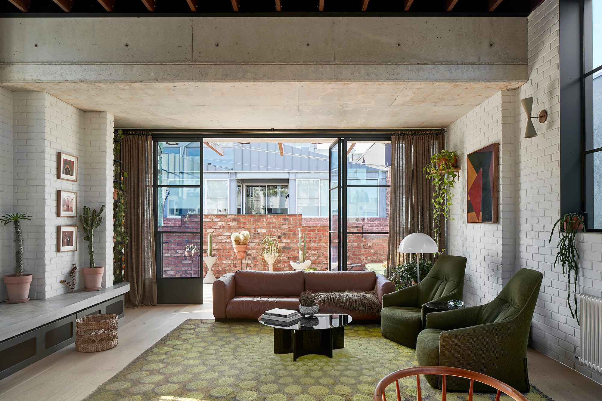 Fabrica by mcmahon and nerlich. Photography by Shannon McGrath. Open plan living space with concrete roof and white brick walls. Green armchairs and brown leather couch. Floor-to-ceiling doors open onto outdoor terrace with red brick wall. 