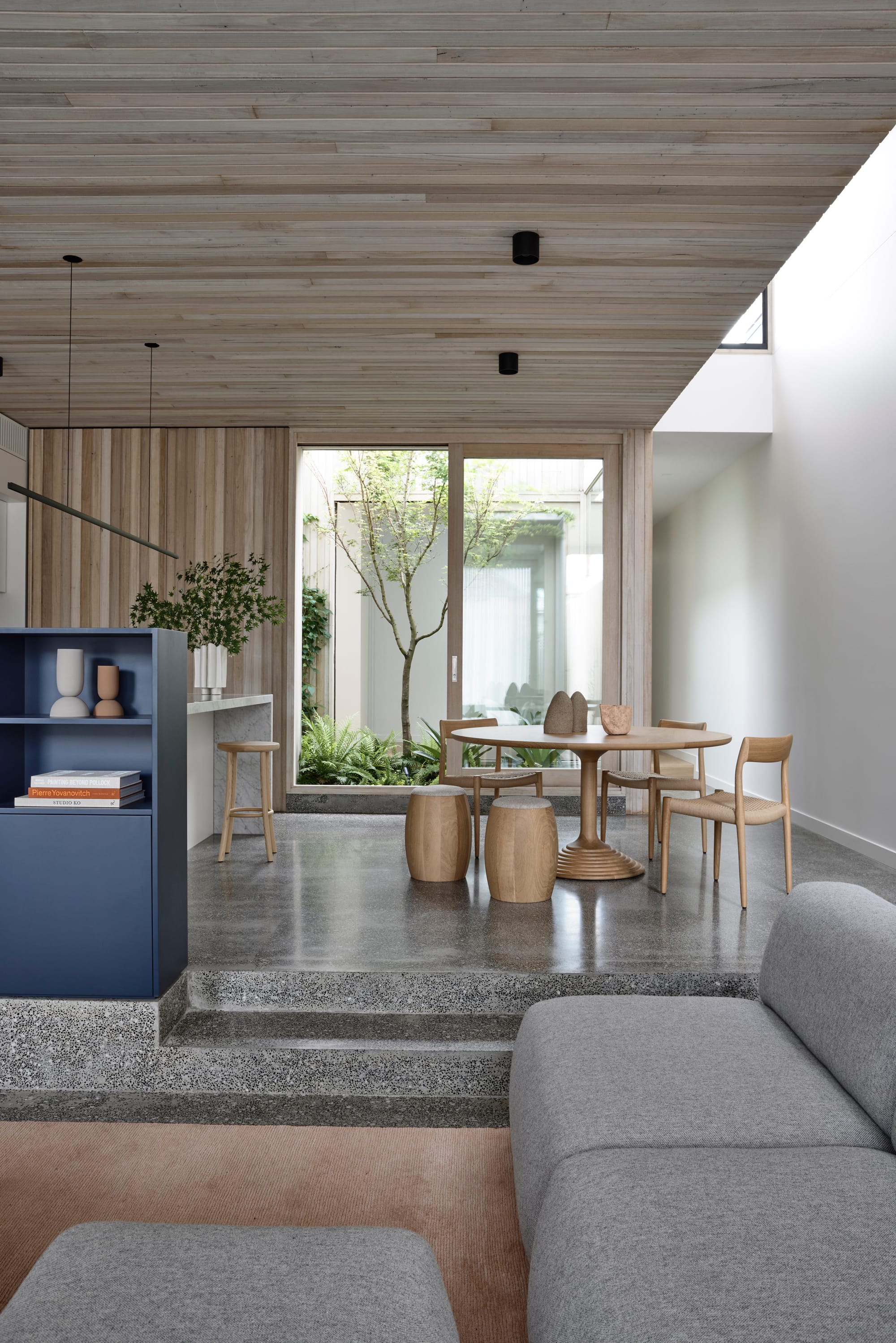 Silvertop House by Tom Robertson Photography. Photography by Derek Swalwell. Polished and speckled concrete raised kitchen and dining area leading away from sunken lounge with grey couch. Round timber dining table. 