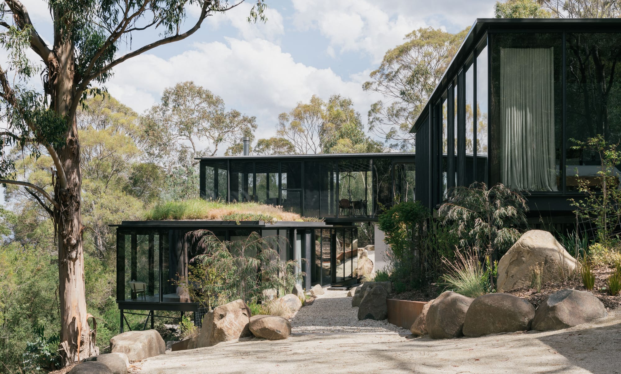 Taroona House by Archier. Photography by Thurston Empson. Residential home constructed of dark framed floor-to-ceiling glass windows. Three distinct rectangular structures arranged in native bushland with stone pathway leading towards. 