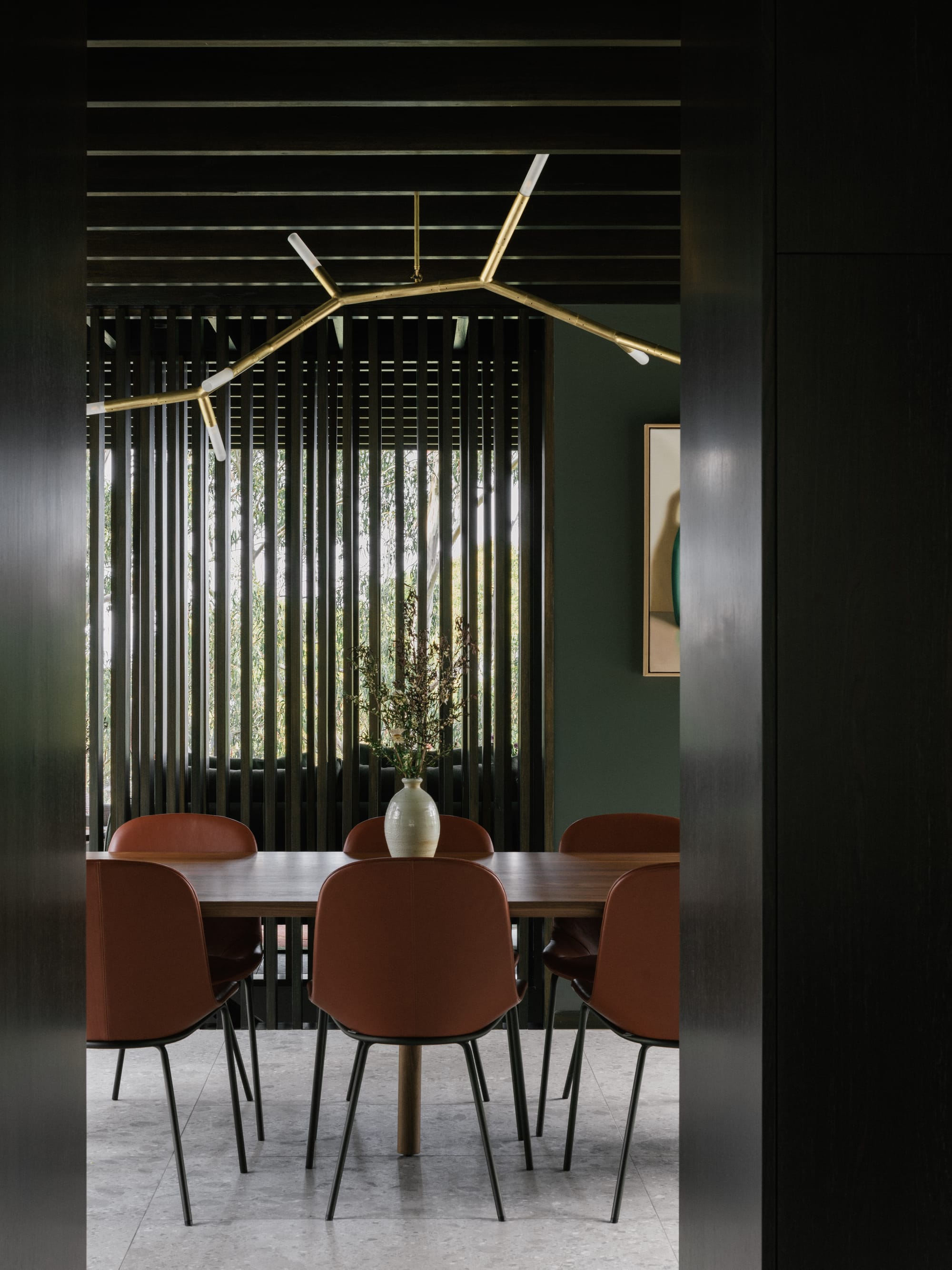 Taroona House by Archier. Photography by Thurston Empson. Dark doorway looking into dining space with timber table and orange chairs. Dark green walls with timber dividing panel. Gold abstract light hanging from ceiling. 