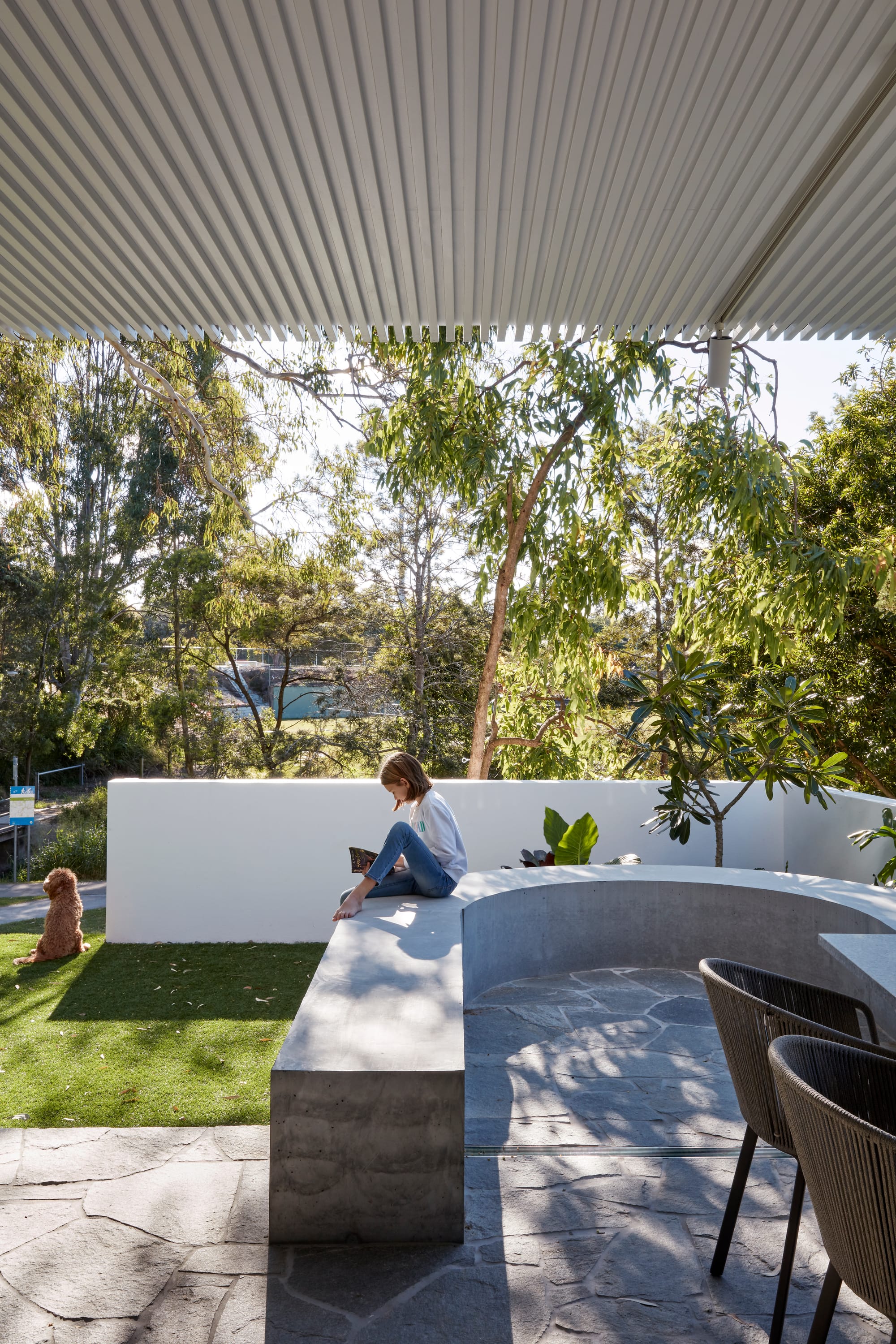 Uxbridge House by Tim Stewart Architects. Photography by Christopher Frederick Jones. Garden with curved concrete half wall enclosing paved dining space. Green grass and tall trees overlooking park. 