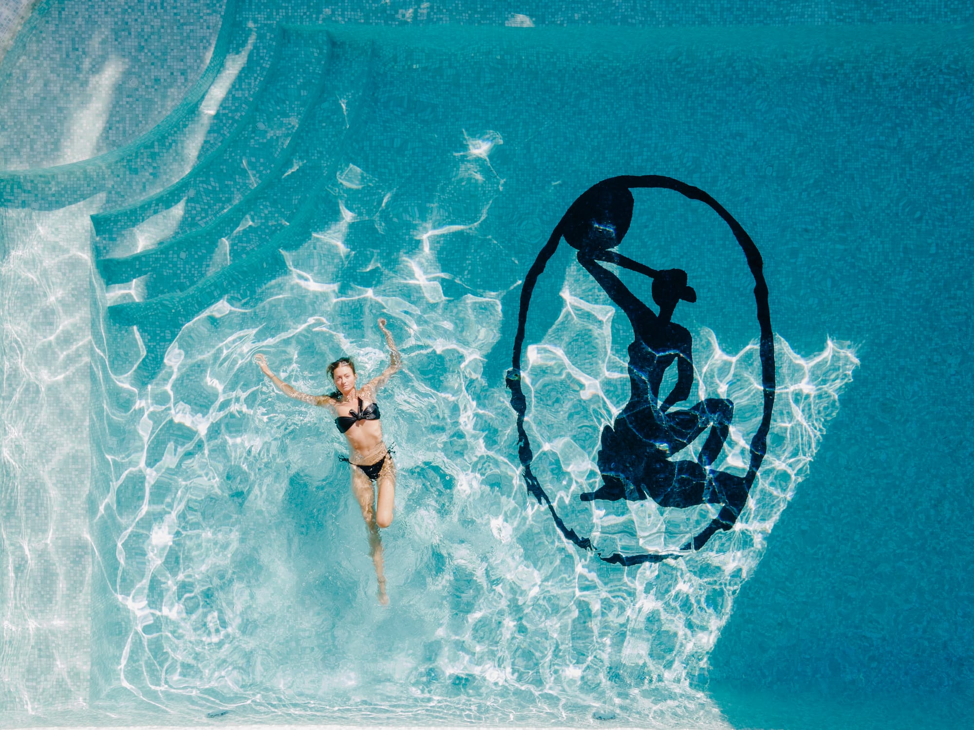 Basq House. Photography by Elise Hassey. Birds eye shot of woman in black bikini floating on her back in blue tiled pool with black logo embedded into pool floor. 