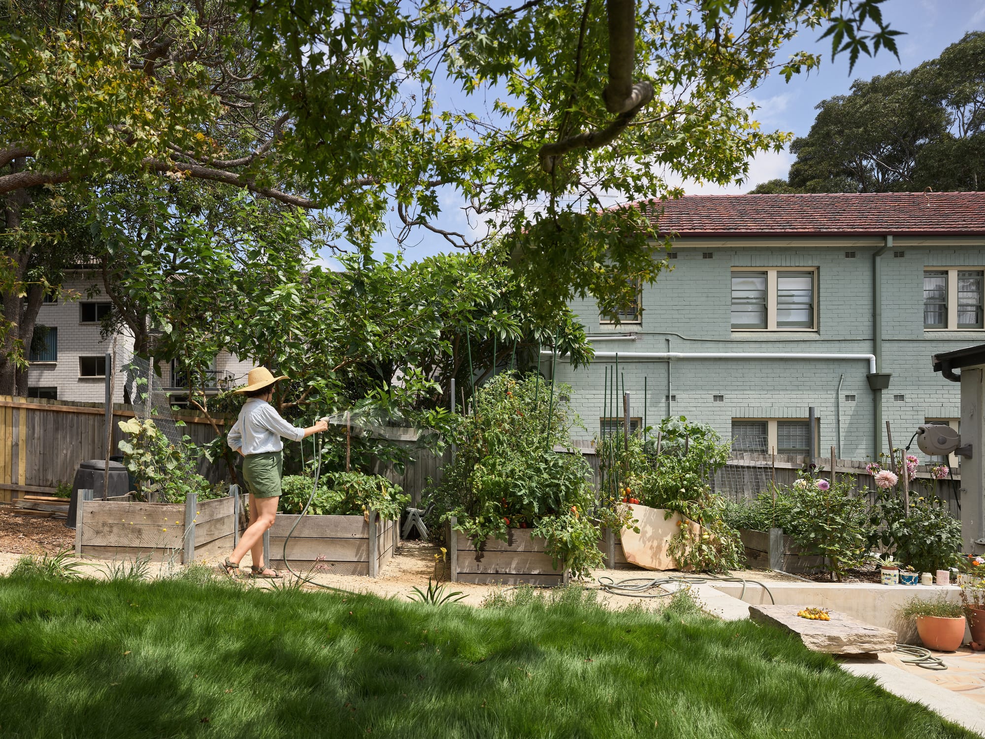 Coconut Crab by Alexander Symes Architect. Photography by Barton Taylor Photography. Vegetable patch being watered by woman in a hat. Long green grass in foreground, double storey teal building in background.
