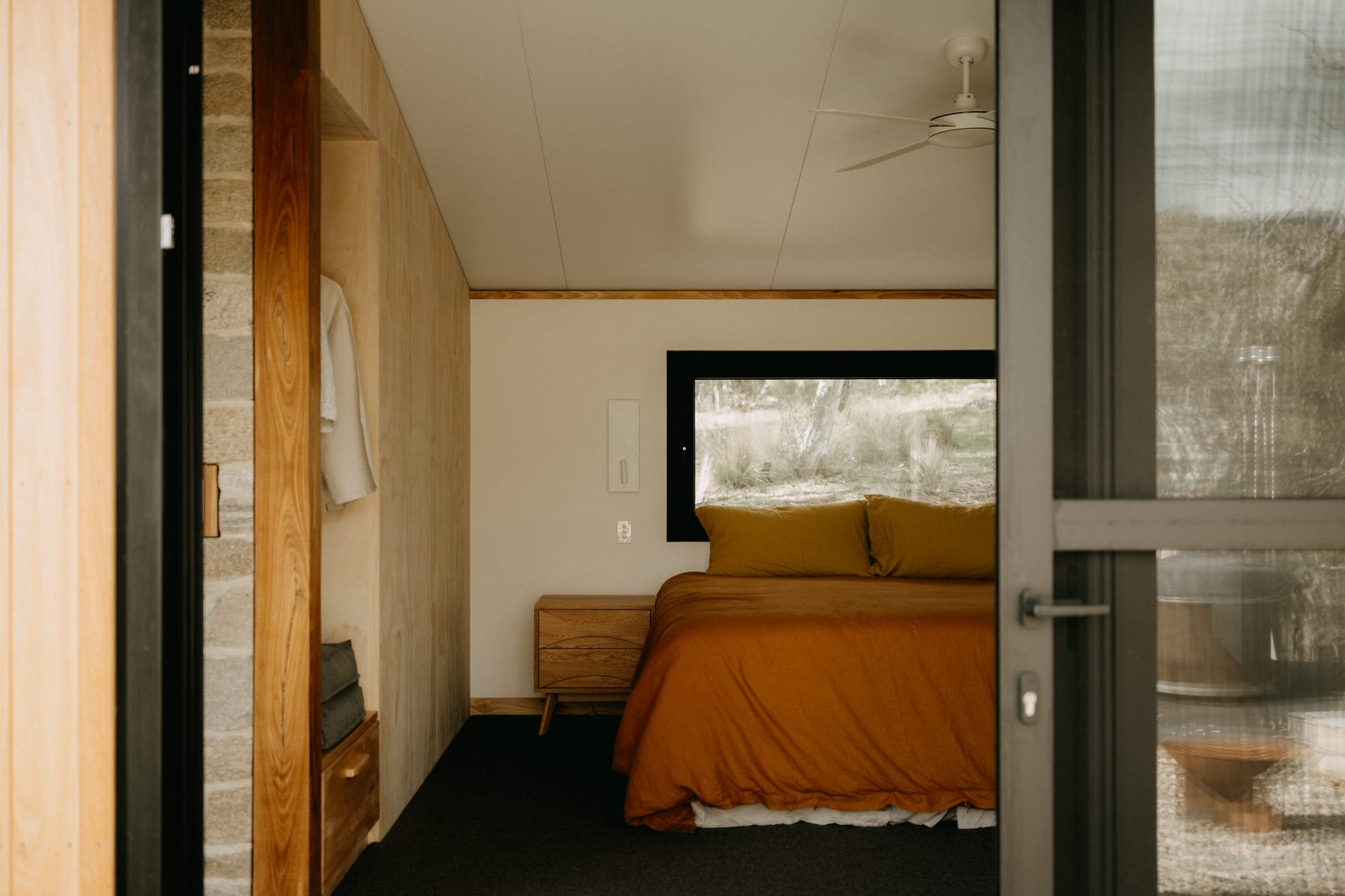 Crafters Eco-Cabins. Image copyright of Crafters. Bedroom with warm linen bedcovers, in room with slanted white ceiling and raw plywood wardrobe.