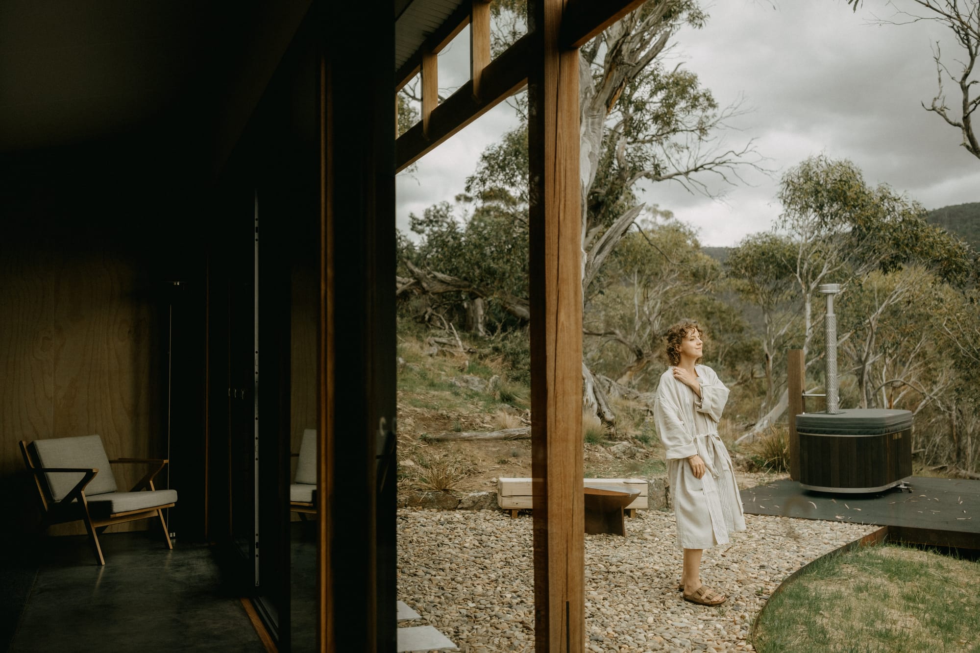 Crafters Eco-Cabins. Image copyright of Crafters. Woman in white robe standing on stone pathway leading from external deck to cabin home. Native Australian trees.