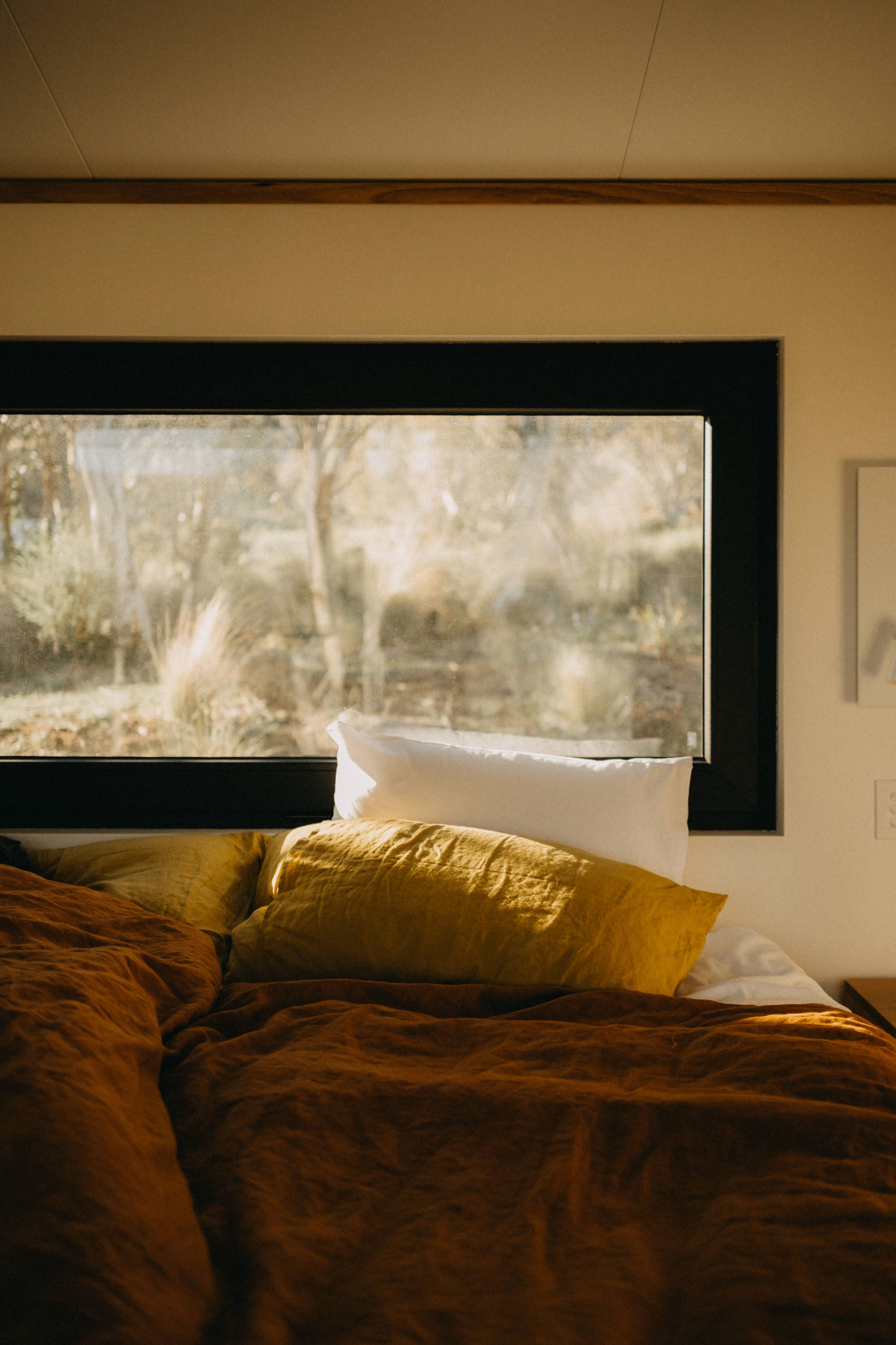 Crafters Eco-Cabins. Image copyright of Crafters. Rust and mustard coloured bed linen in front of black framed landscape window overlooking Australian bushland. 