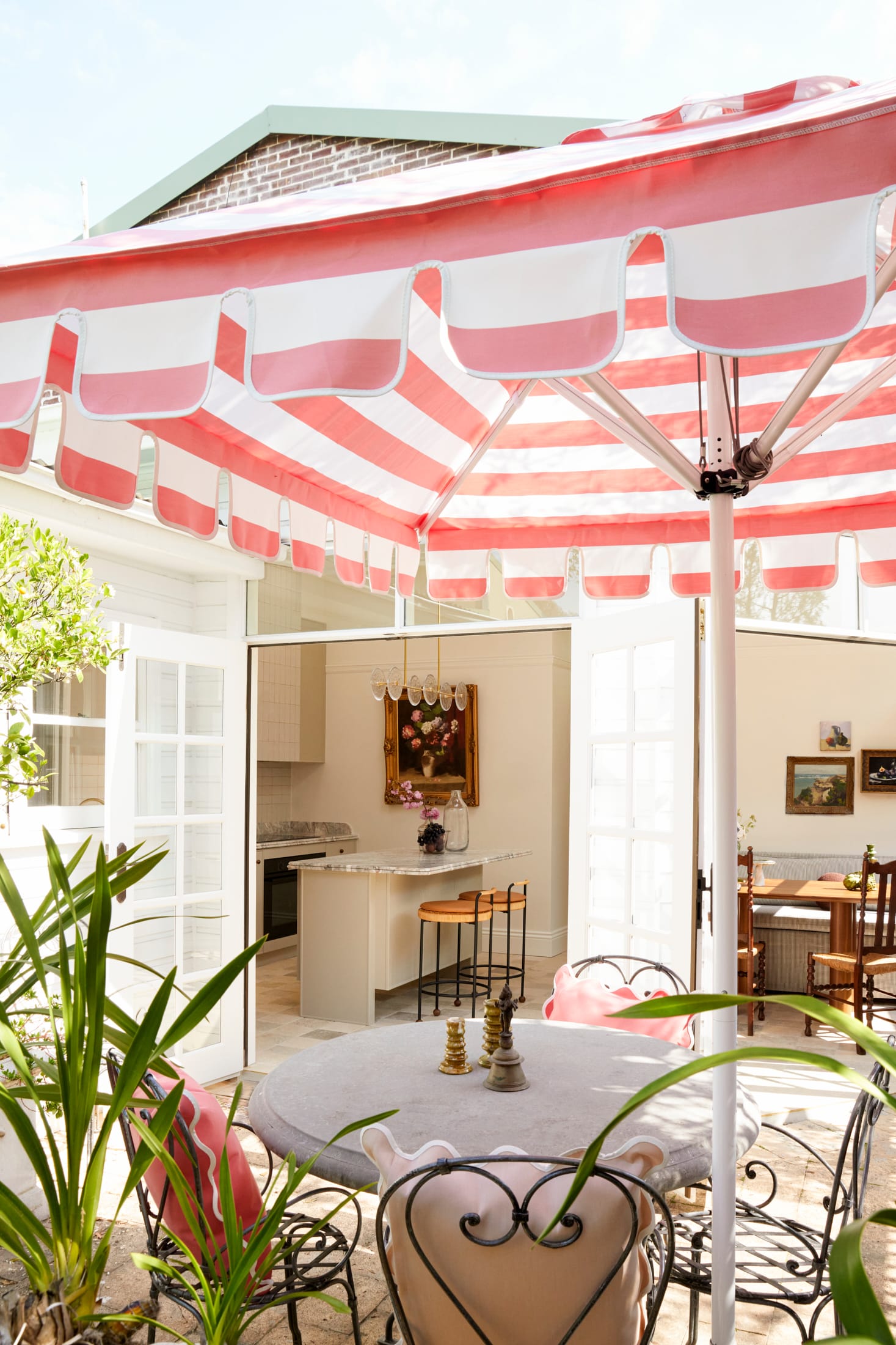 Drummoyne House by studio BARBARA. Photography by Jacqui Turk.  Outdoor dining setting with large pink and white striped umbrella. Wrought iron vintage dining chairs surrounded by green plants.