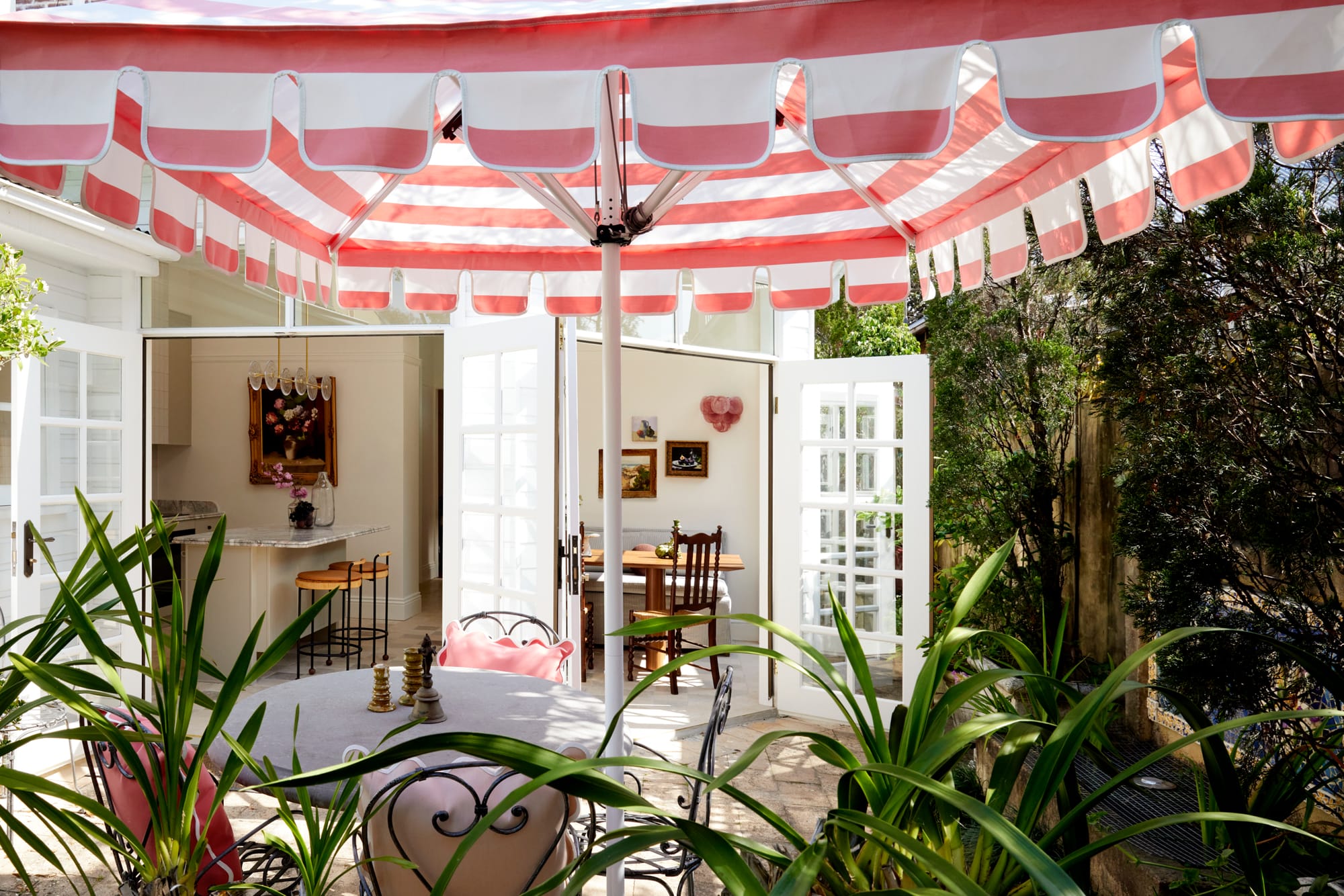 Drummoyne House by studio BARBARA. Photography by Jacqui Turk. Garden terrace with large pink and white umbrella over outdoor wrought iron dining setting. Lush gardens. French doors open onto indoor dining space and kitchen.