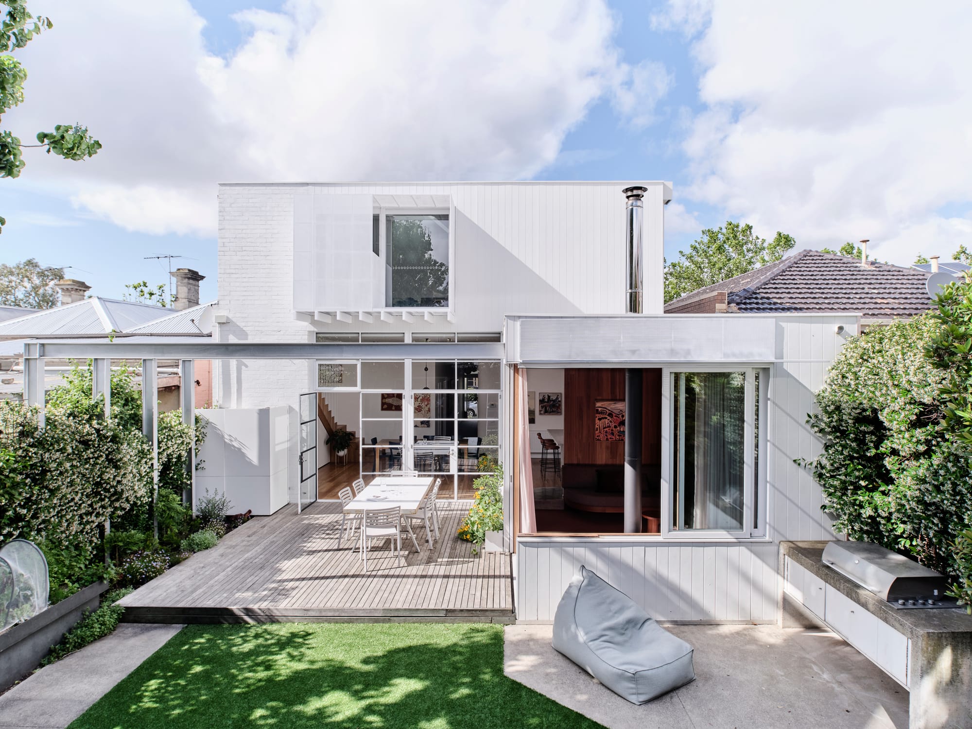 Fitzroy North Residence. Photography by Dean Bradley. Exterior facade of double storey white clad home with pergola and timber deck. Concrete floor outdoor kitchen next to grass.