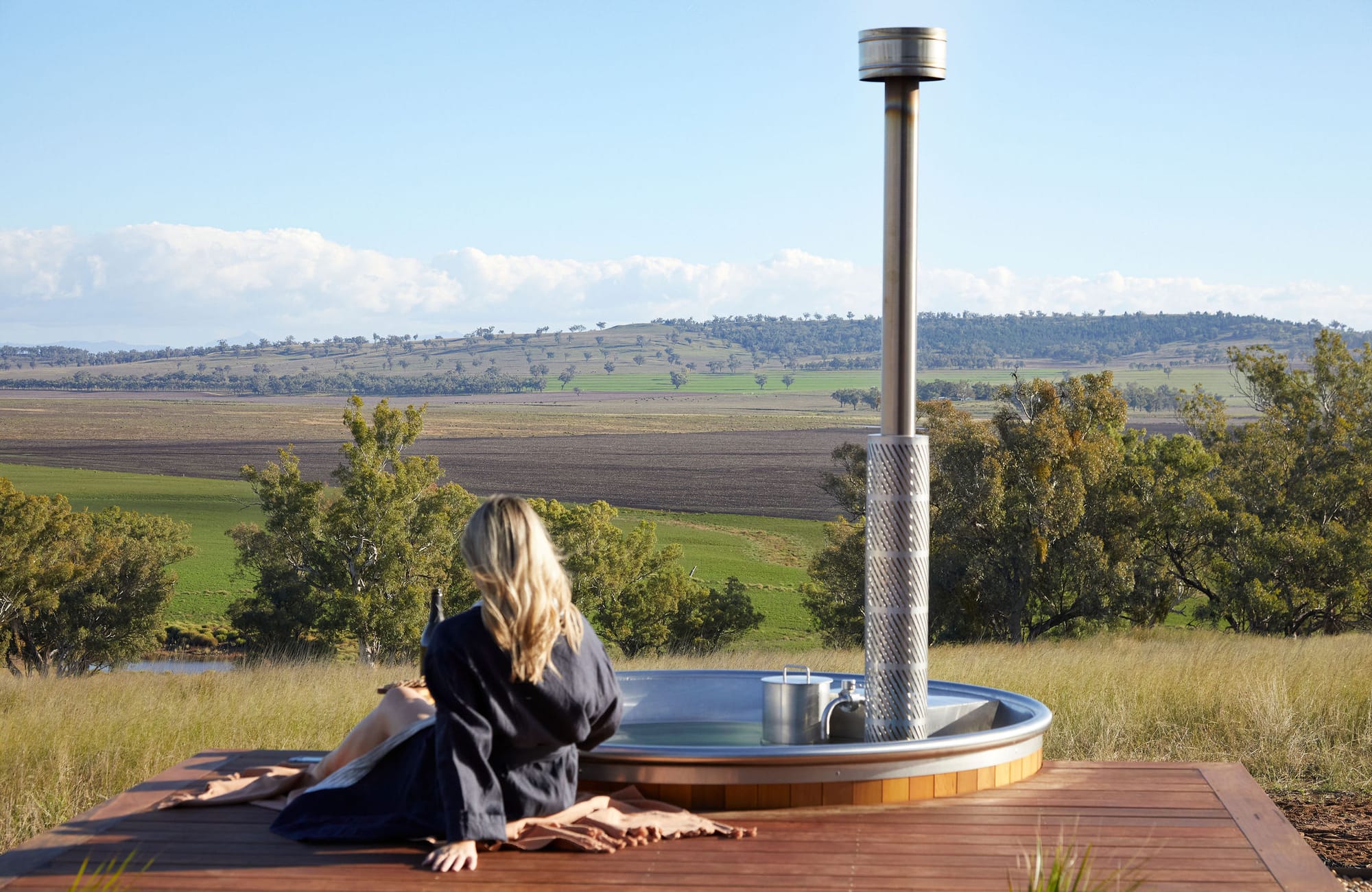 Gilay Estate by Cameron Anderson Architects. Photography by Morning Swim. Woman lounging on timber deck next to sunken hot tub, overlooking views of hilly countryside.