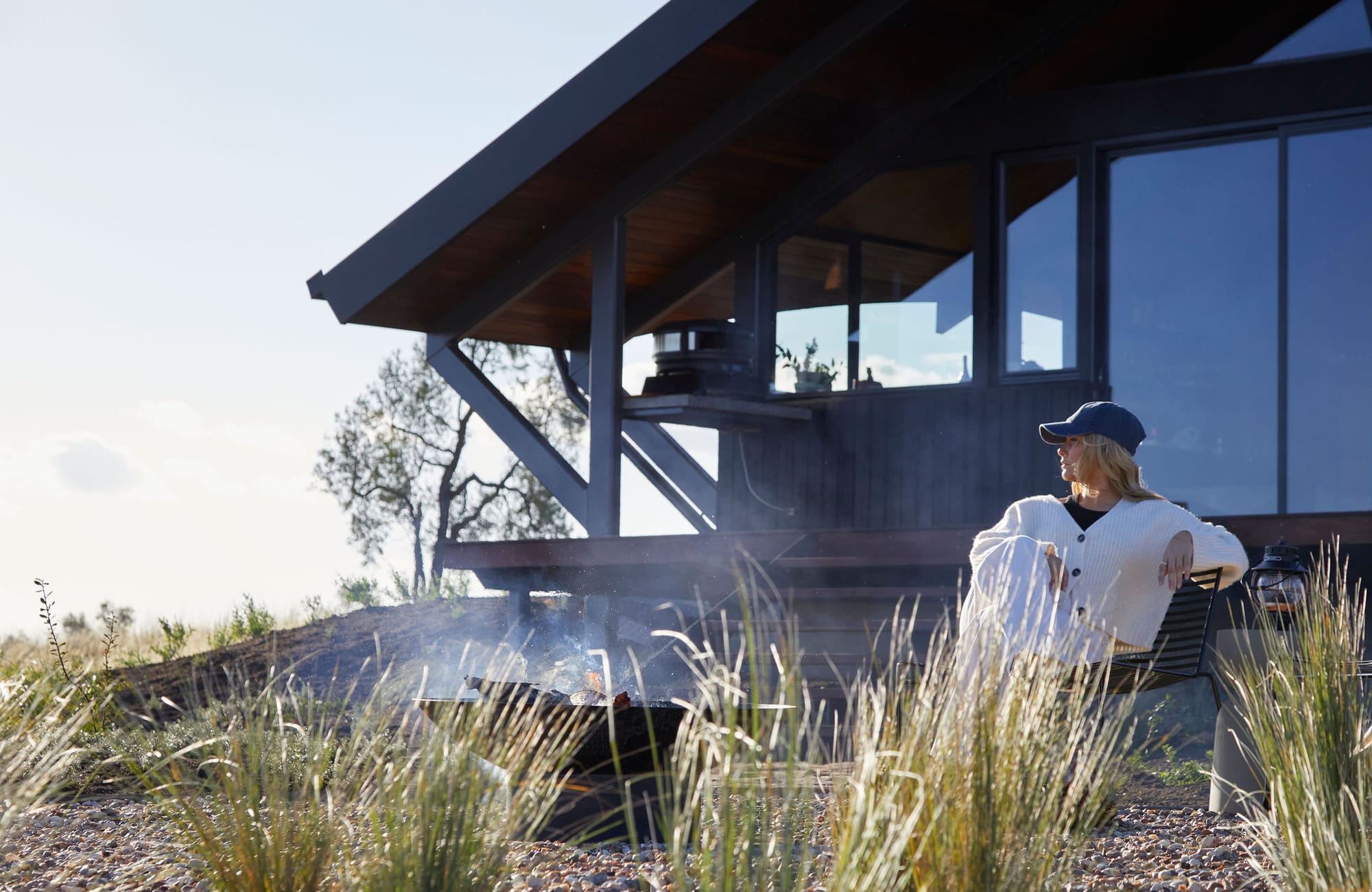 Gilay Estate by Cameron Anderson Architects. Photography by Morning Swim. Woman in white sits on chair next to outdoor firepit in front of black timber clad angled building.