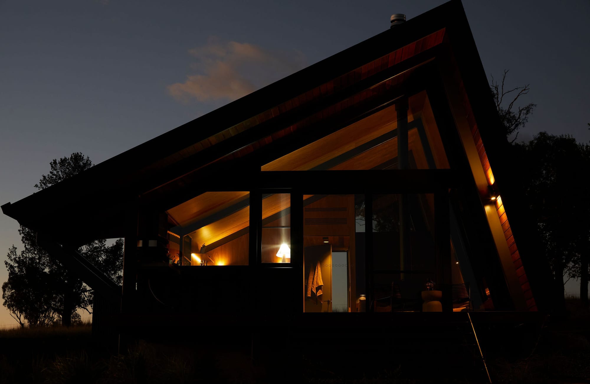 Gilay Estate by Cameron Anderson Architects. Photography by Morning Swim. Angled building lit from within at night. Warm light bouncing against timber interior.