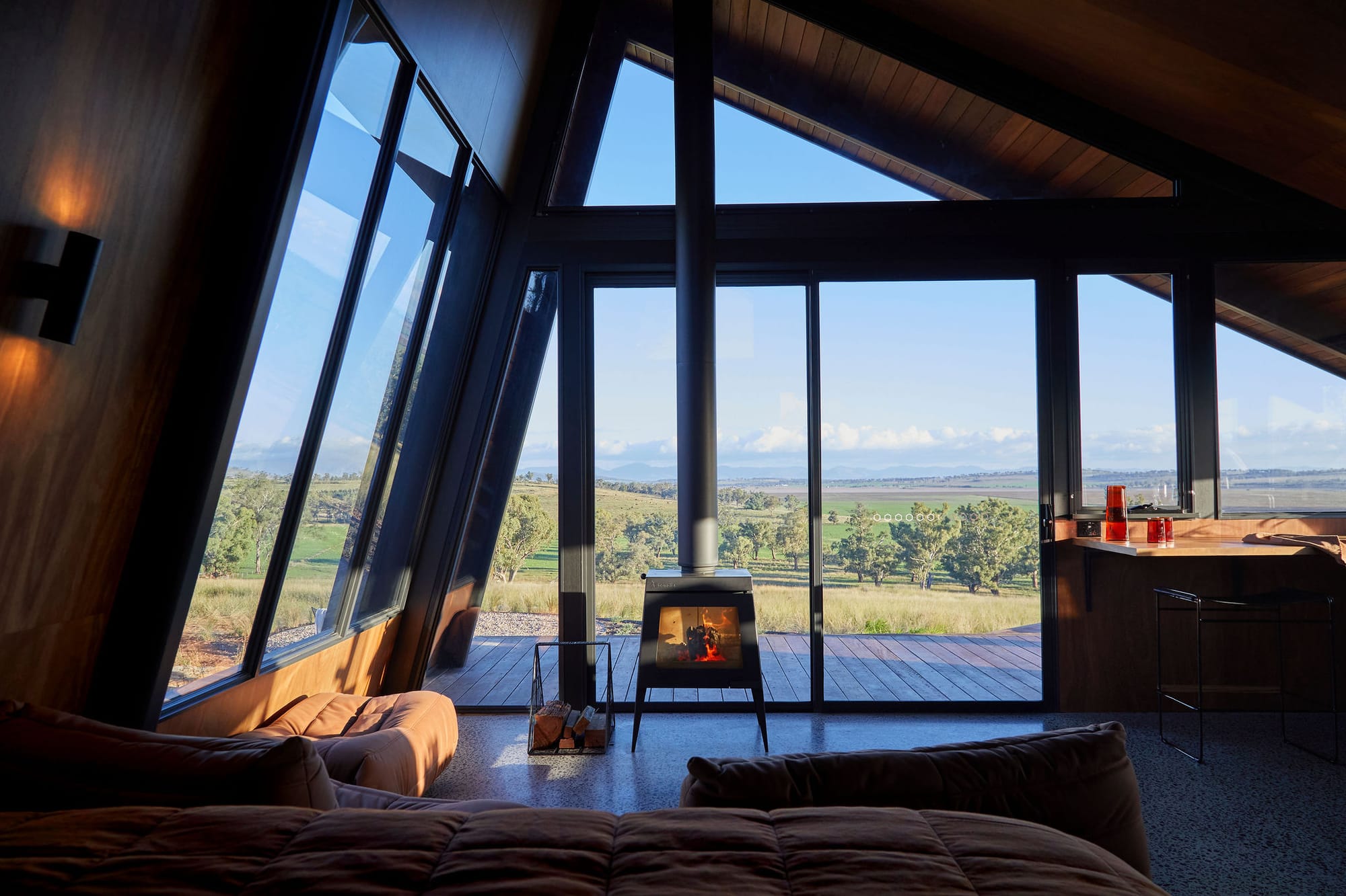 Gilay Estate by Cameron Anderson Architects. Photography by Morning Swim. Angled floor-to-ceiling windows overlooking timber deck and rolling hillside. Metal fireplace in front of window on concrete floor. 