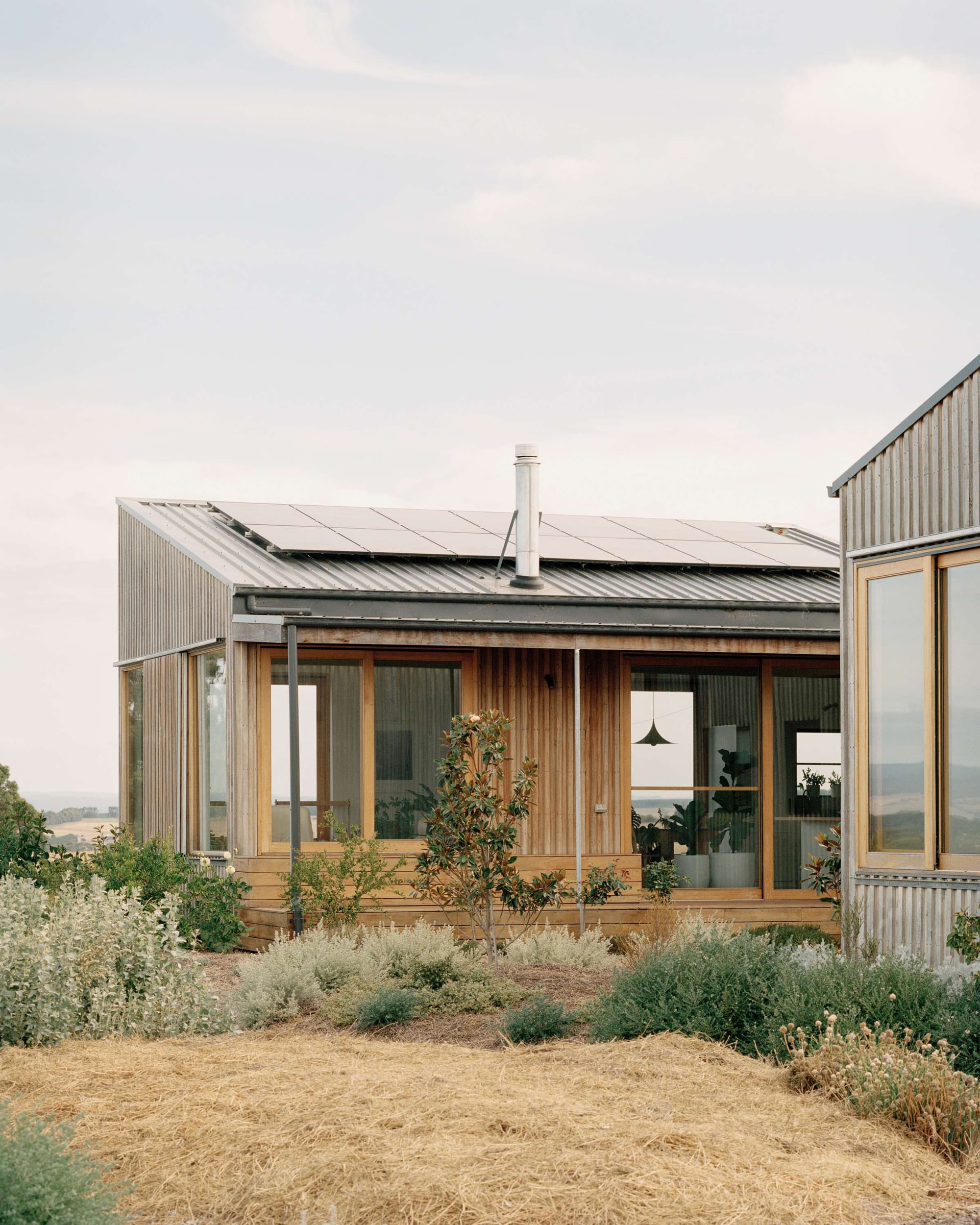 Heather's Off-Grid House by Gardiner Architects. Photography by Rory Gardiner. Facade of timber clad farmhouse with tin roof with solar panels. Dry grass and scattered garden bed in foreground. 