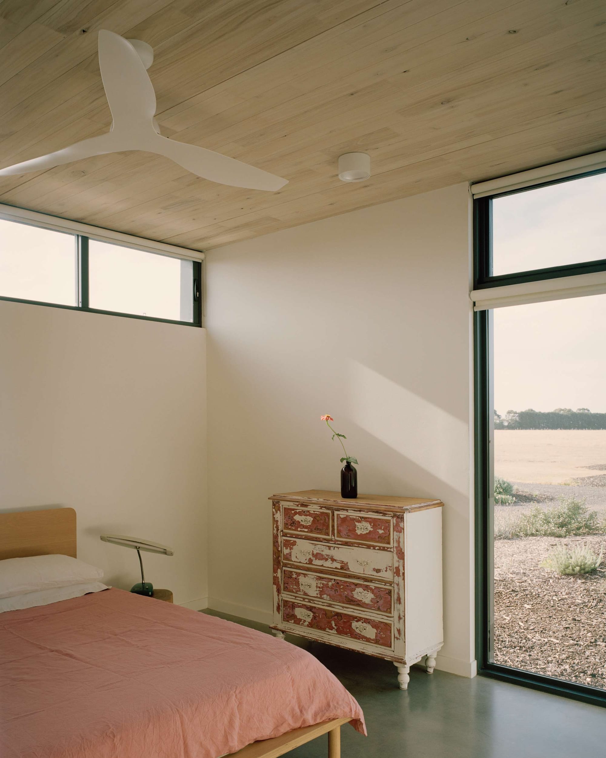 Heather's Off-Grid House by Gardiner Architects. Photography by Rory Gardiner. Bedroom with polished concrete floors, white walls, timber roof and large window overlooking barren land. Bed with pink linen and white and pink dresser distressed in shabby-chic style.