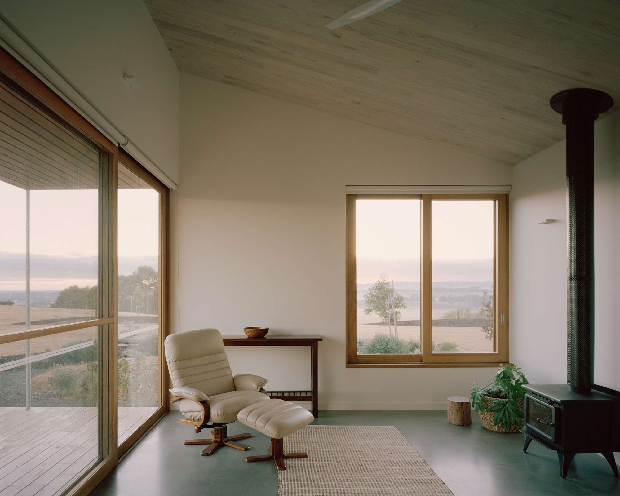 Heather's Off-Grid House by Gardiner Architects. Photography by Rory Gardiner. Sitting room with white walls, angled timber ceiling, polished concrete floors, single white armchair and traditional black fireplace. 