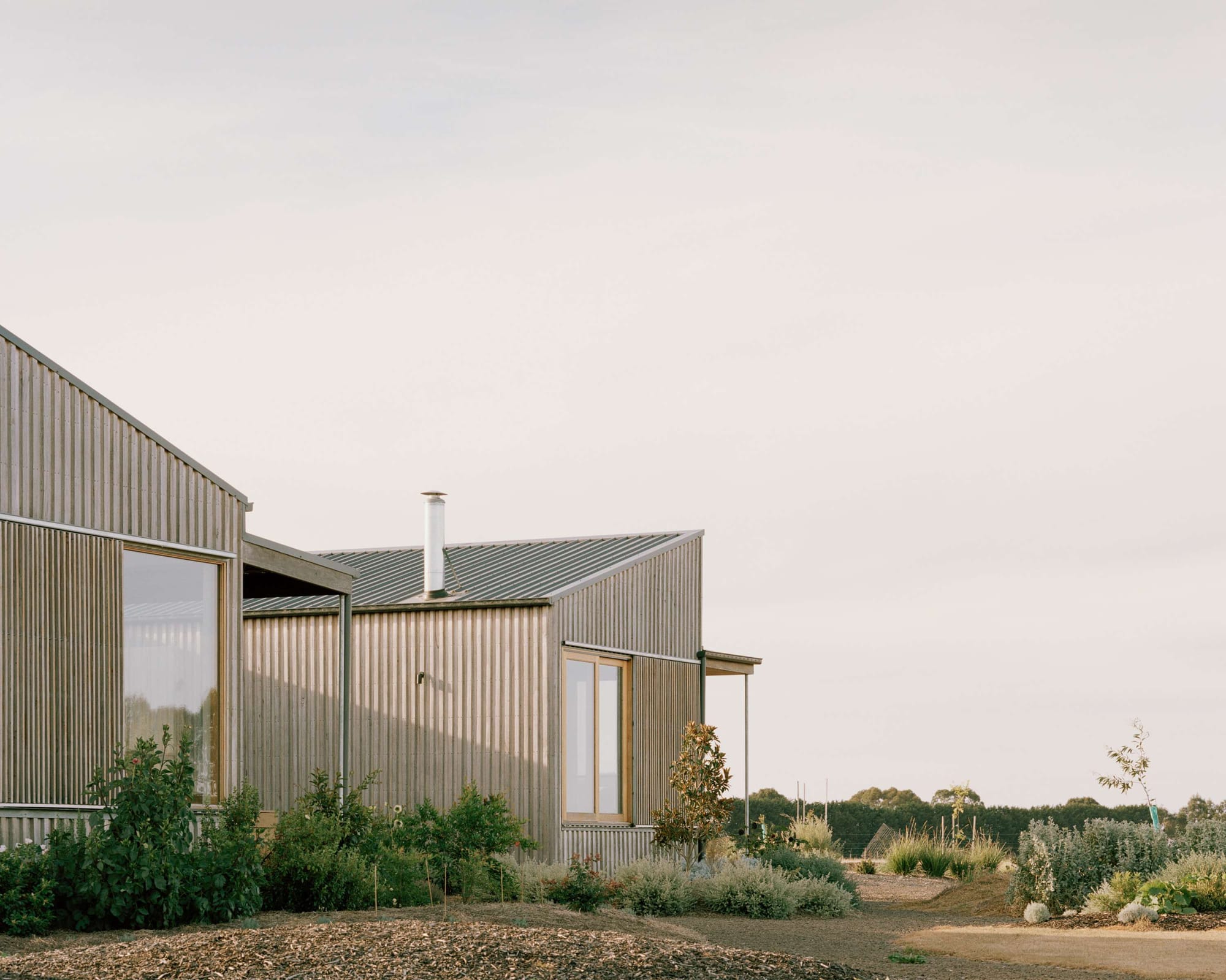 Heather's Off-Grid House by Gardiner Architects. Photography by Rory Gardiner. Facade of timber clad farmhouse surrounded by dry and barren paddock with scattered garden beds and native plants. 