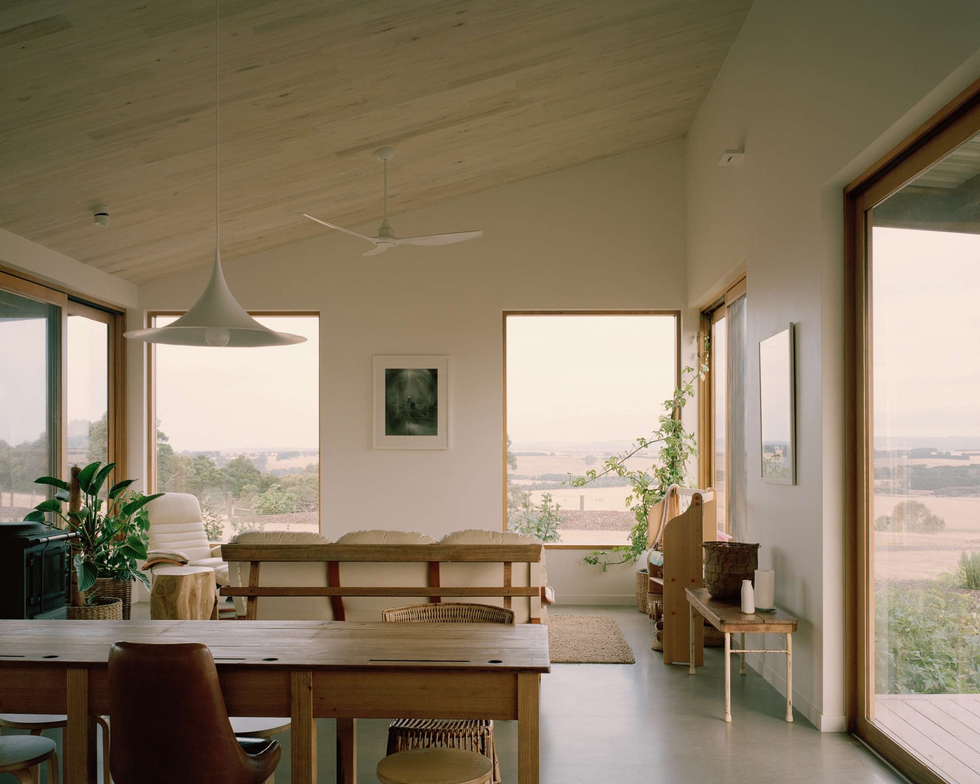 Heather's Off-Grid House by Gardiner Architects. Photography by Rory Gardiner. Dining and living space with high angled white walls and timber ceiling and polished concrete floor. Dry rolling paddocks visible out of timber framed windows. 