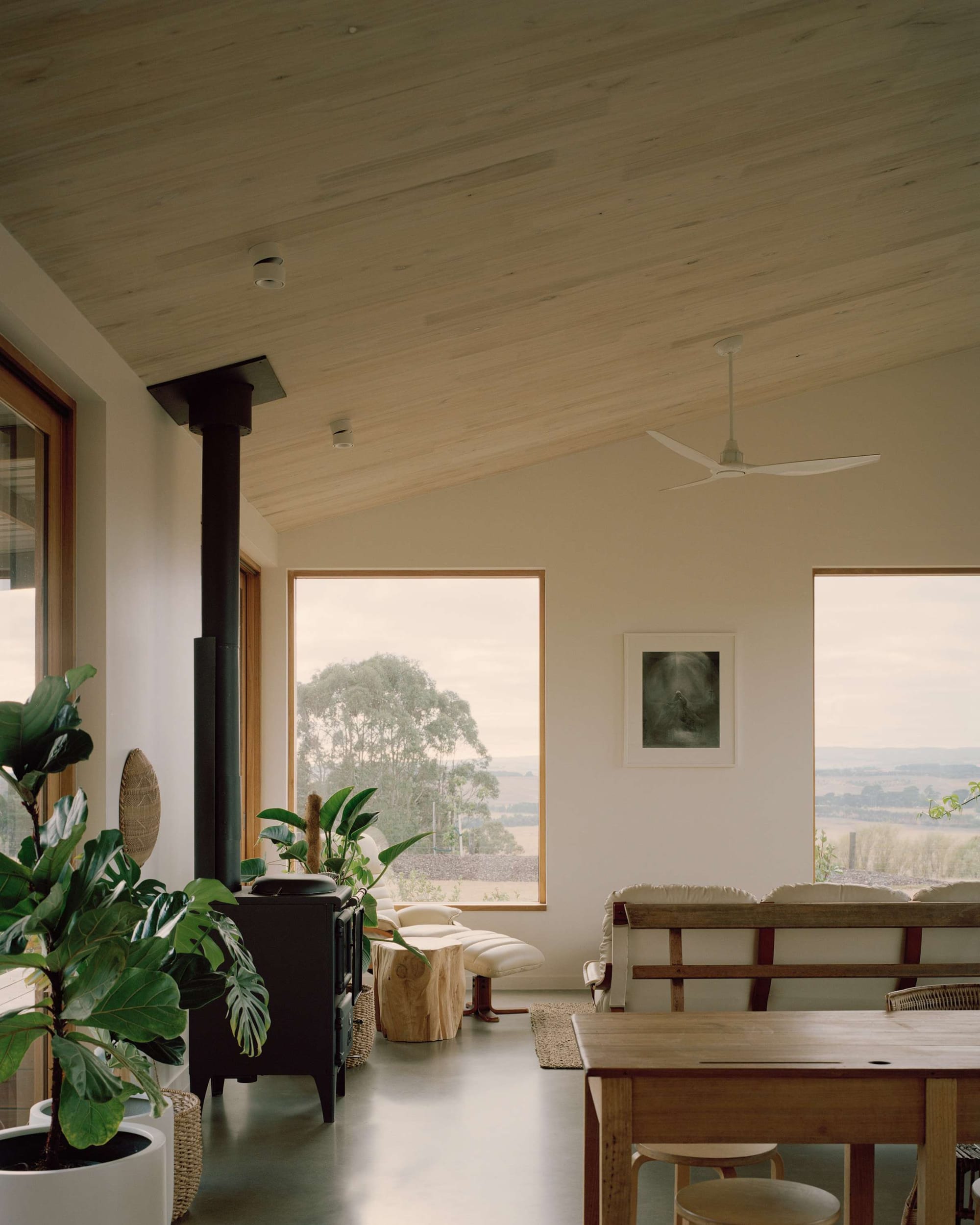 Heather's Off-Grid House by Gardiner Architects. Photography by Rory Gardiner. Living and dining space with timber framed windows overlooking rolling paddocks in background. Black fireplace on polished concrete flooring. 