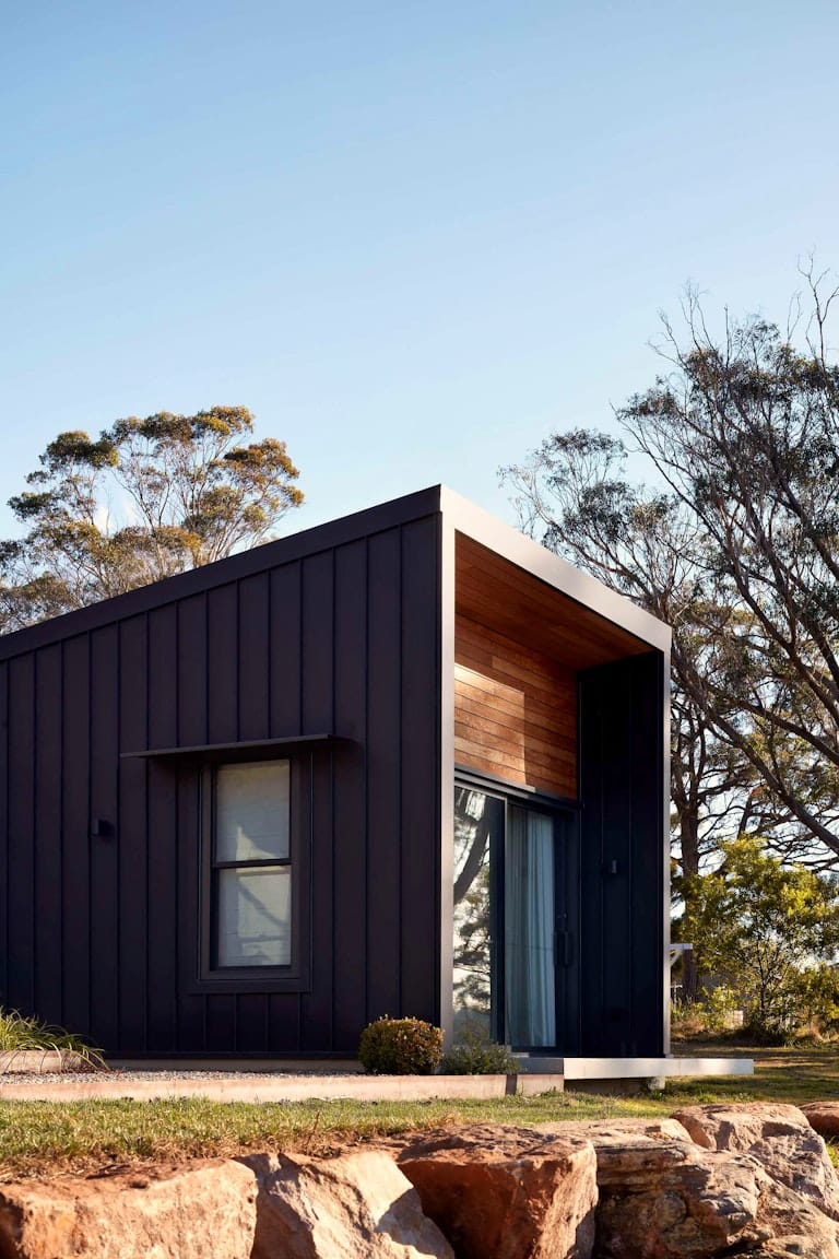 House Woodlands by AO Design Studio. Photograohy by Luc Remond. Exterior of residential home with black and timber clad. Rocks in foreground, native plants in background. 