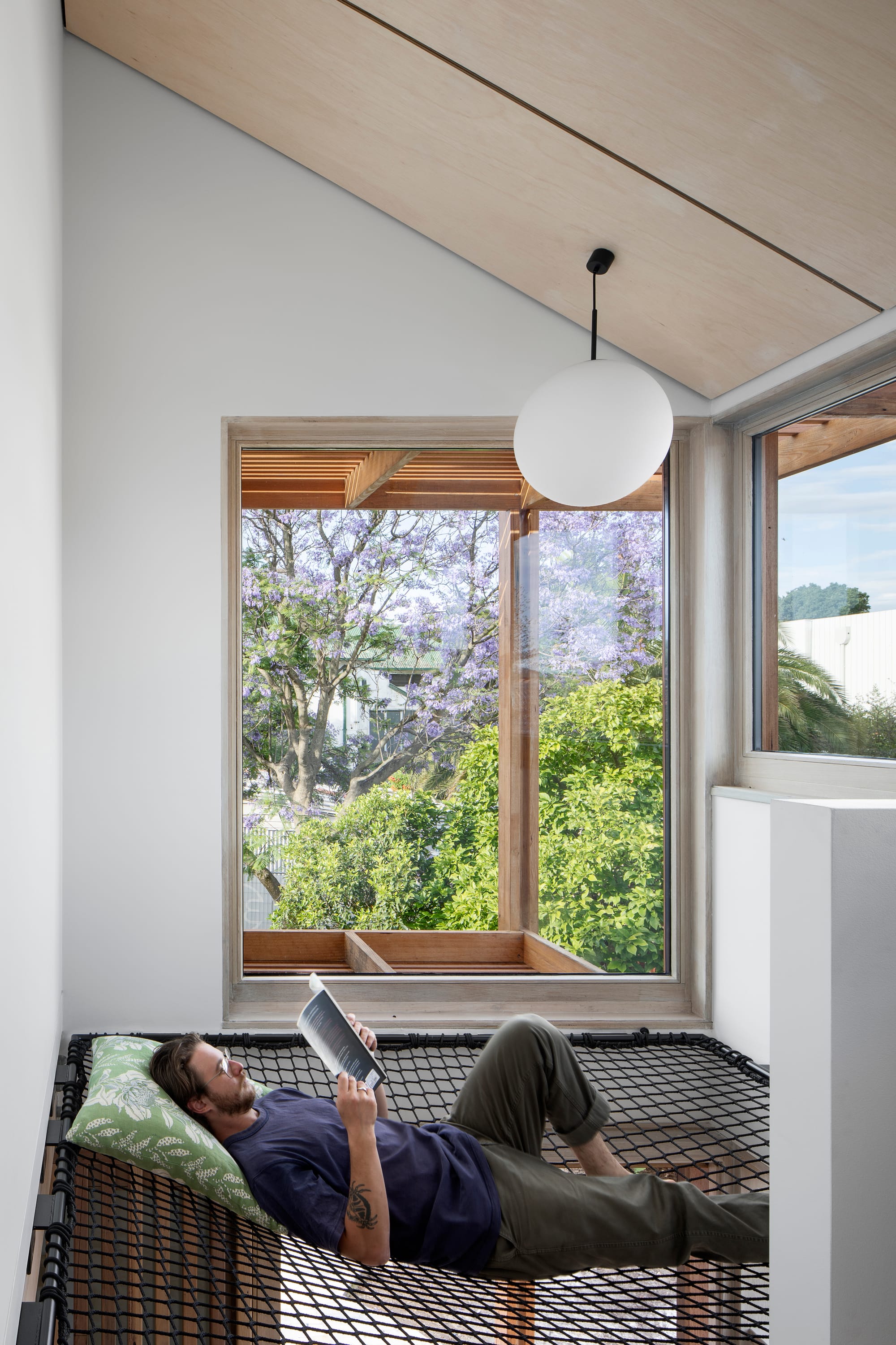 A man reading a book on a net seat with a large window showing the tops of treetops