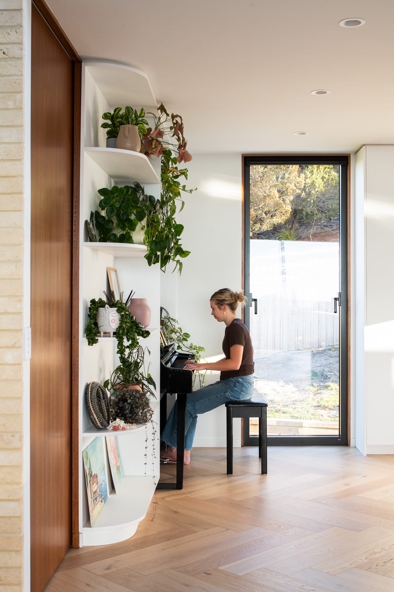 Mortlock Timber Series: The Milldam. Photography by Anjie Blair. Girl playing piano behind floor-to-ceiling white curved cabinetry with plants on it. Herringbone timber floors and windows overlooking bushland. 