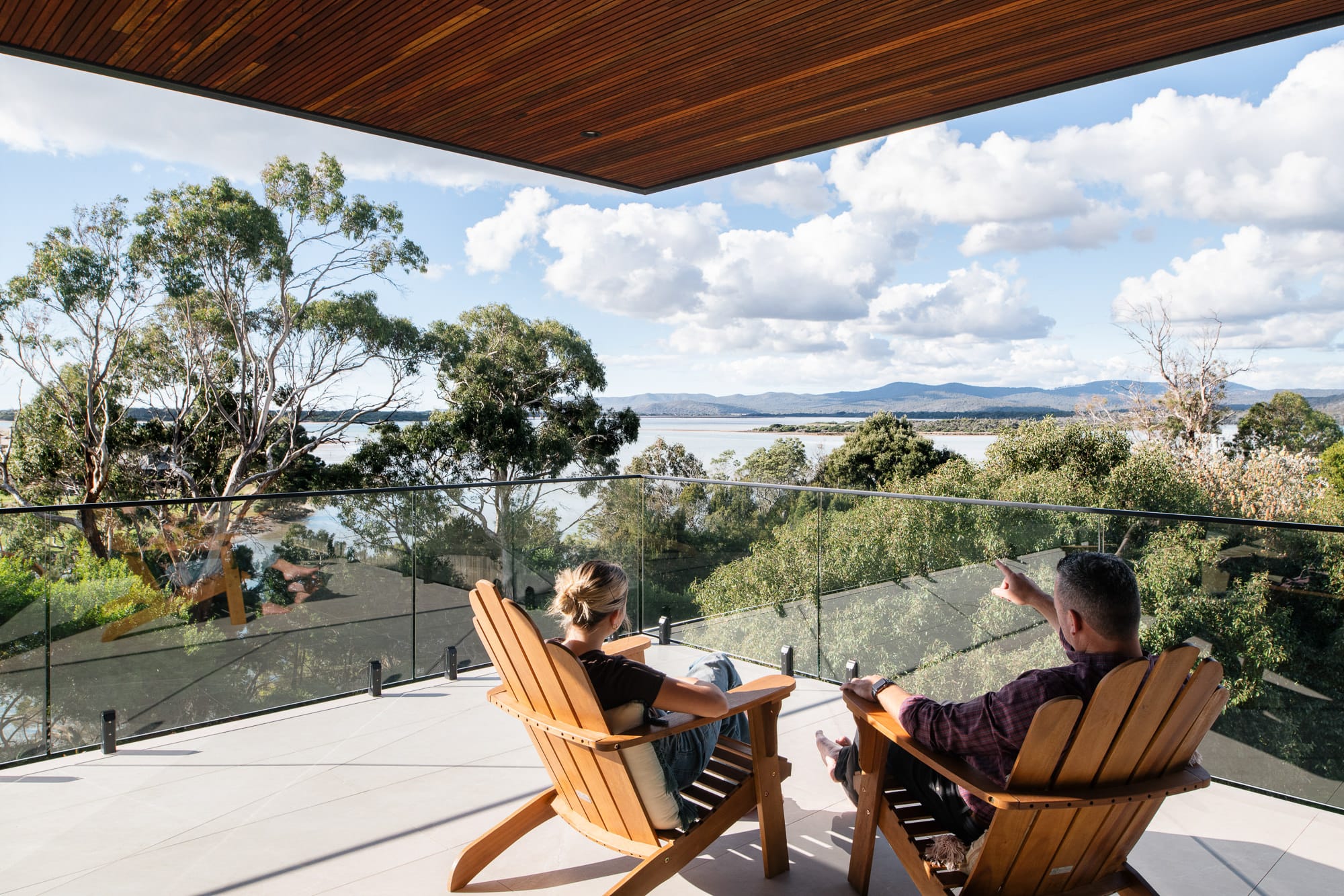 Mortlock Timber Series: The Milldam. Photography by Anjie Blair. Two people sit in timber patio chairs on balcony overlooking estuary and native Australian trees. 